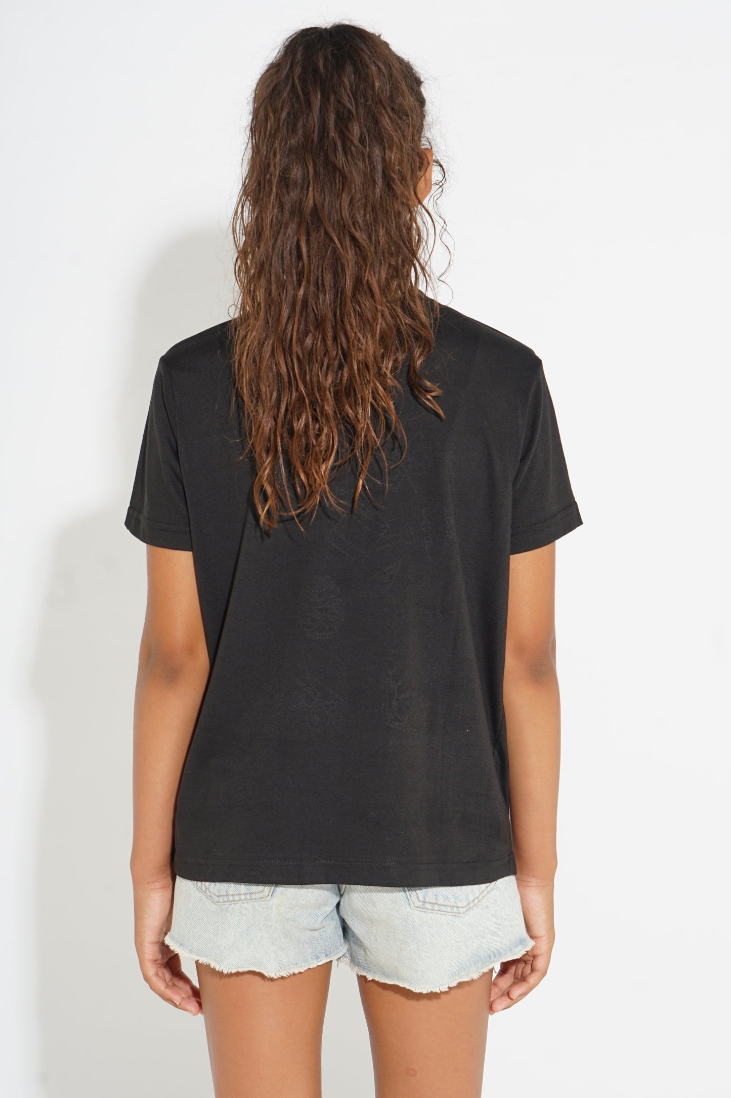 A woman viewed from the back wearing a faded black short-sleeve t-shirt and light-wash denim shorts against a white studio background.
