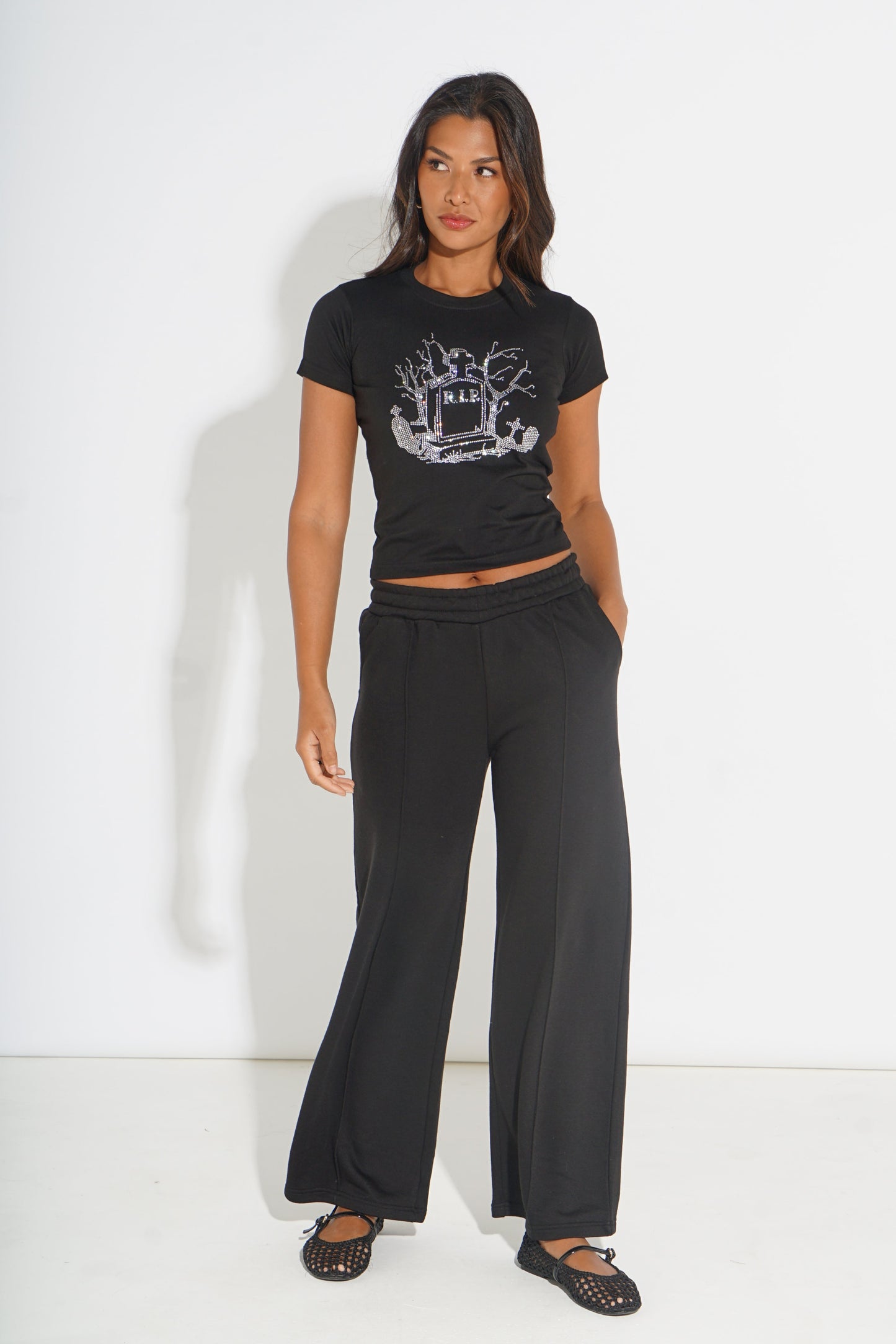 A woman modeling a black short-sleeve crop top with a silver graphic print and matching black wide-leg pants, standing against a plain white studio background.