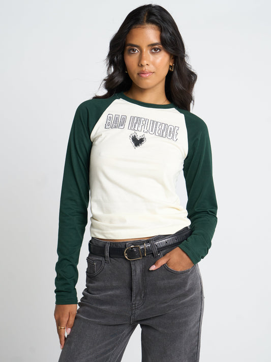 A woman modeling a long-sleeve raglan baseball tee with a cream body and dark green sleeves, featuring 'BAD INFLUENCE' text and a rooster graphic on the chest, paired with dark gray jeans and a black belt against a light gray studio background.