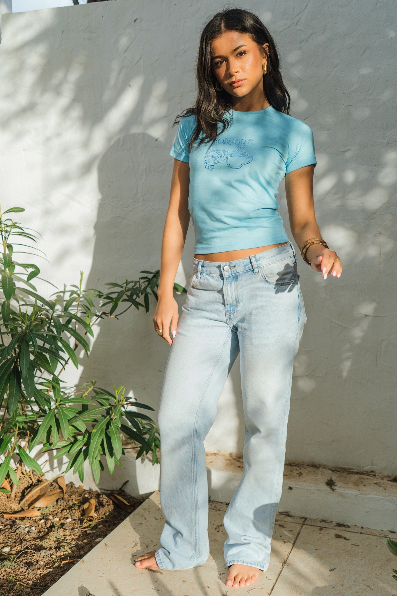 Woman wearing a blue tie-dye shirt and light blue jeans standing against a white wall with a plant to the left.