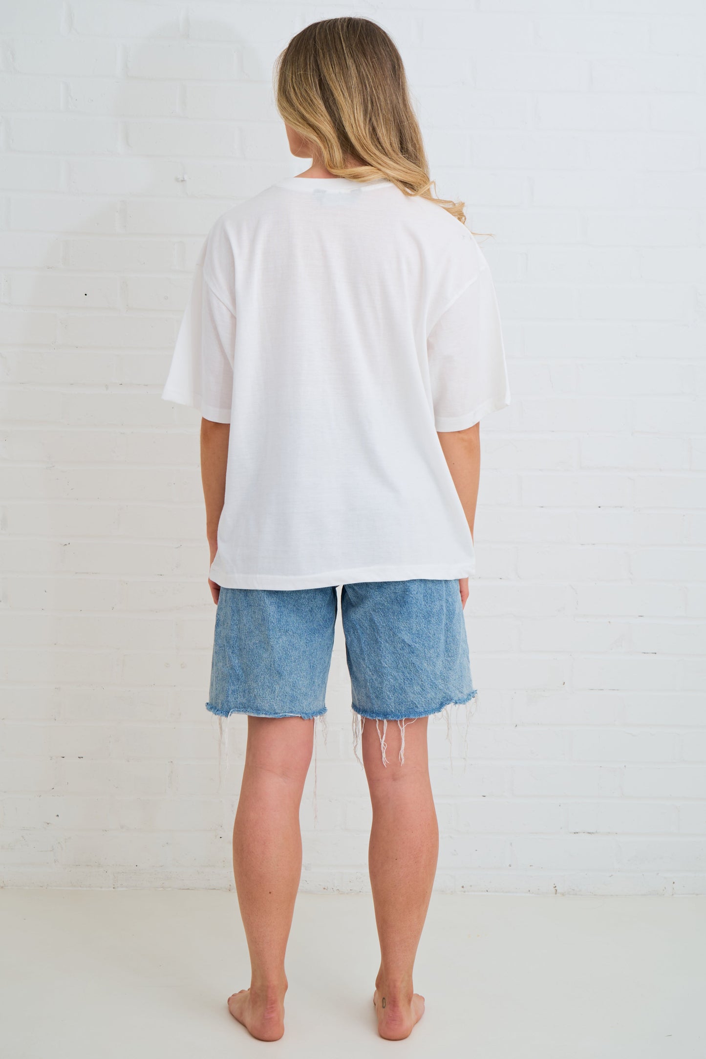 Person wearing a white oversized t-shirt and denim shorts against a white brick wall.