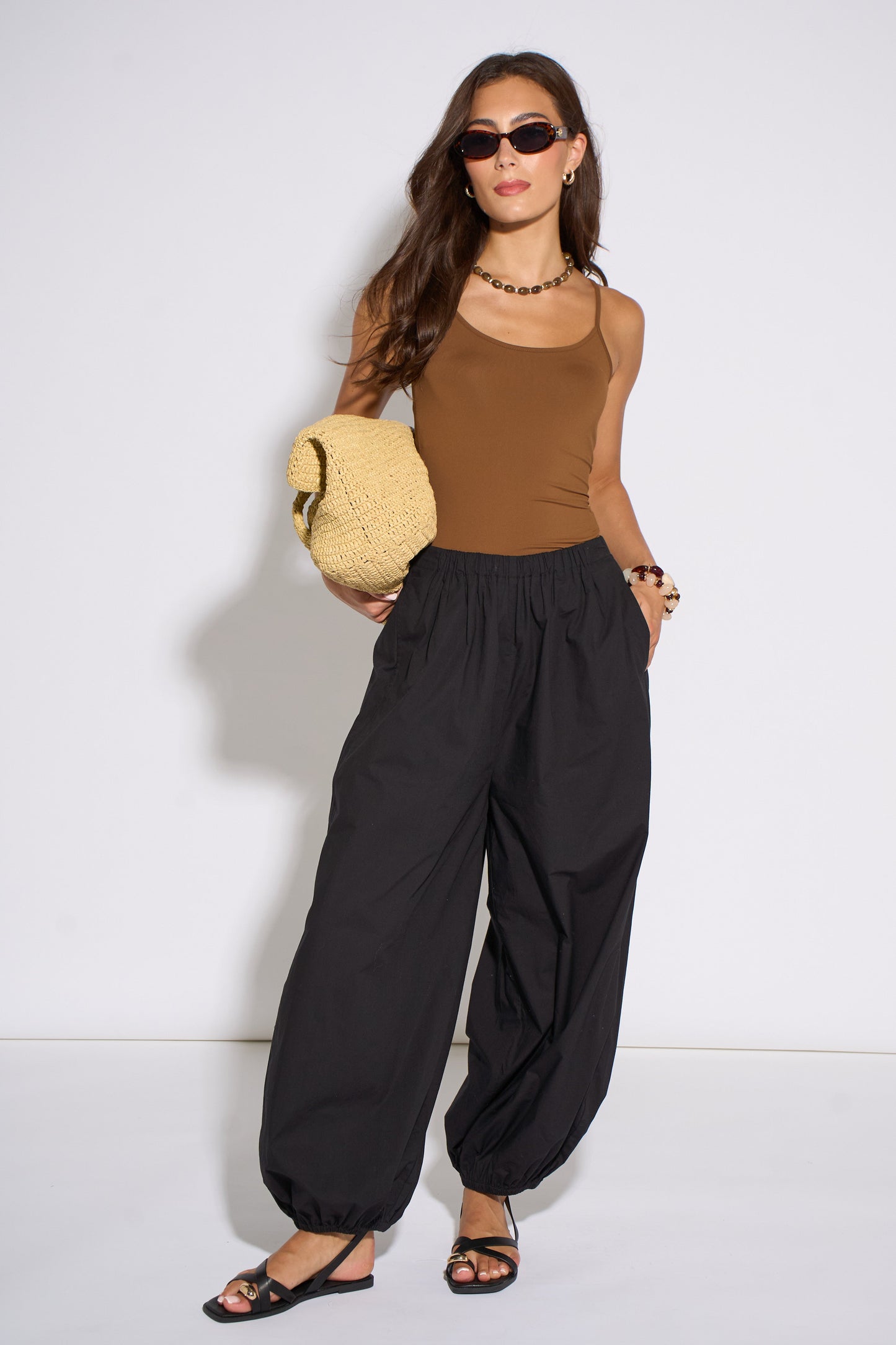 Woman wearing a brown top and black pants holding a beige hat against a white background