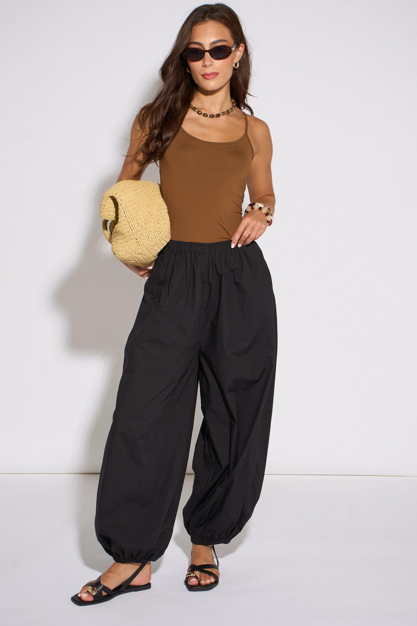 Woman wearing a brown top and black pants holding a beige bag against a white background