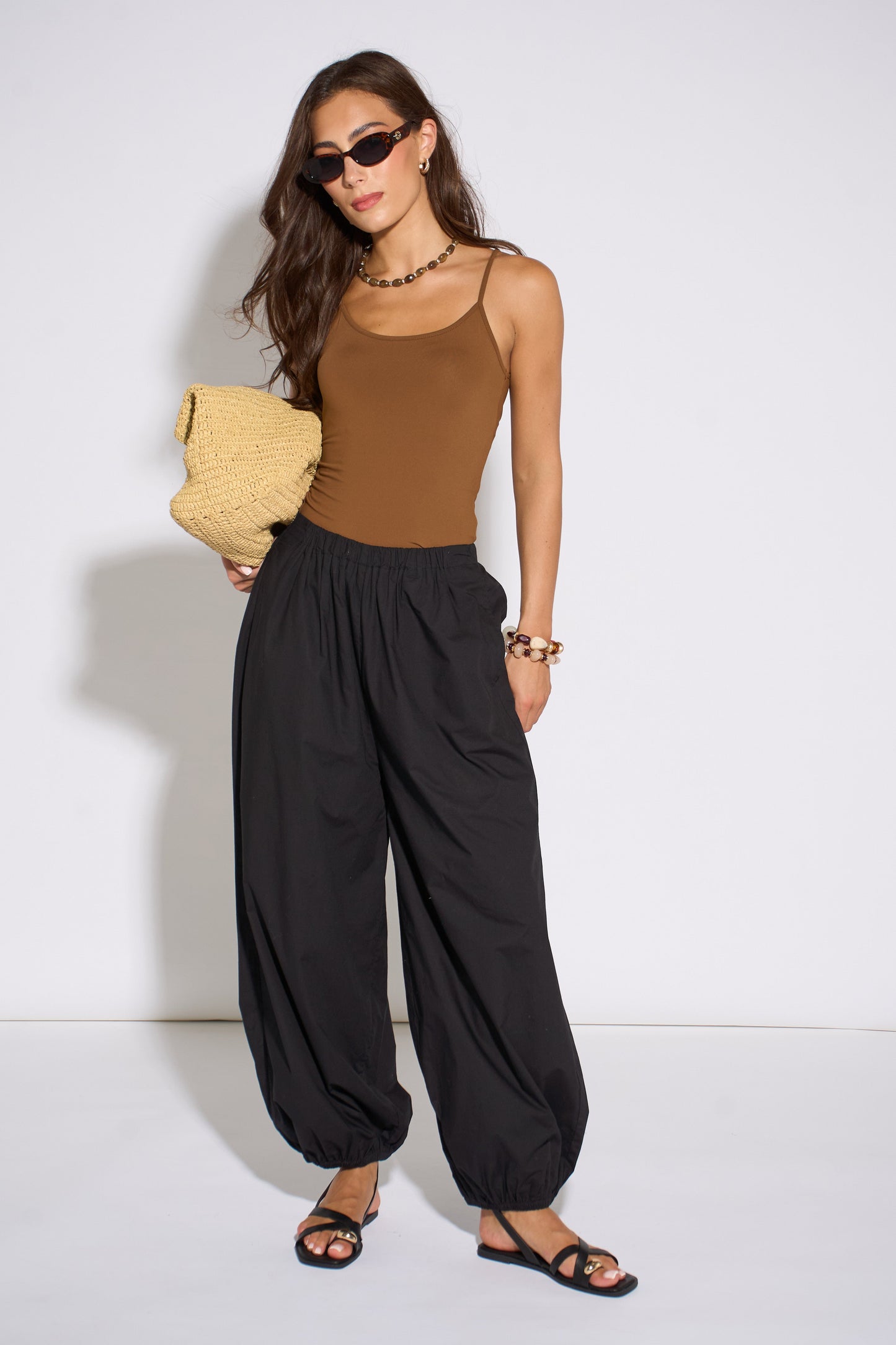 Woman wearing a brown top and black high-waisted pants holding a yellow clutch against a white background