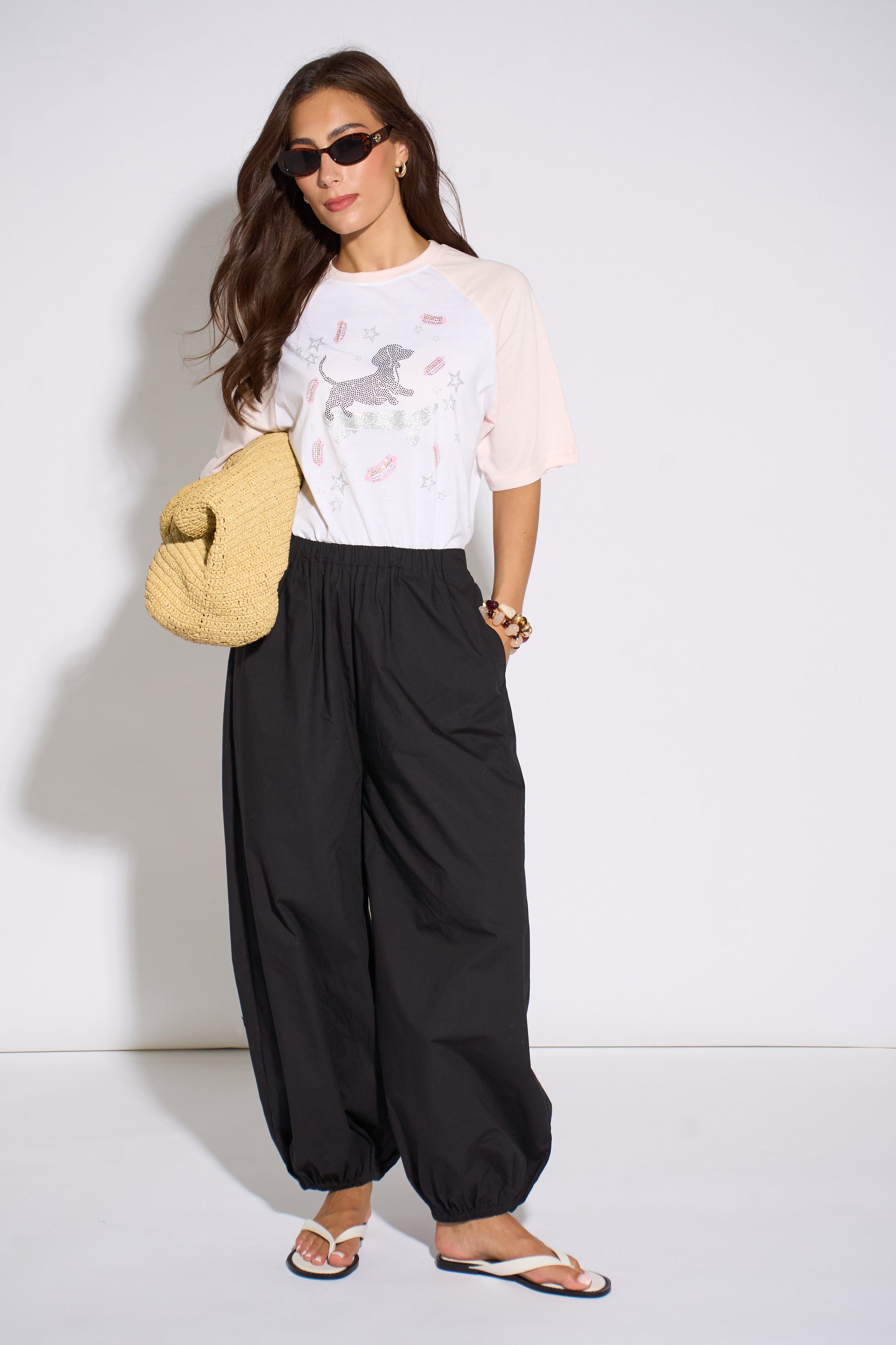 Woman wearing a white t-shirt with a graphic design and black wide-leg pants, holding a yellow bag against a white background.