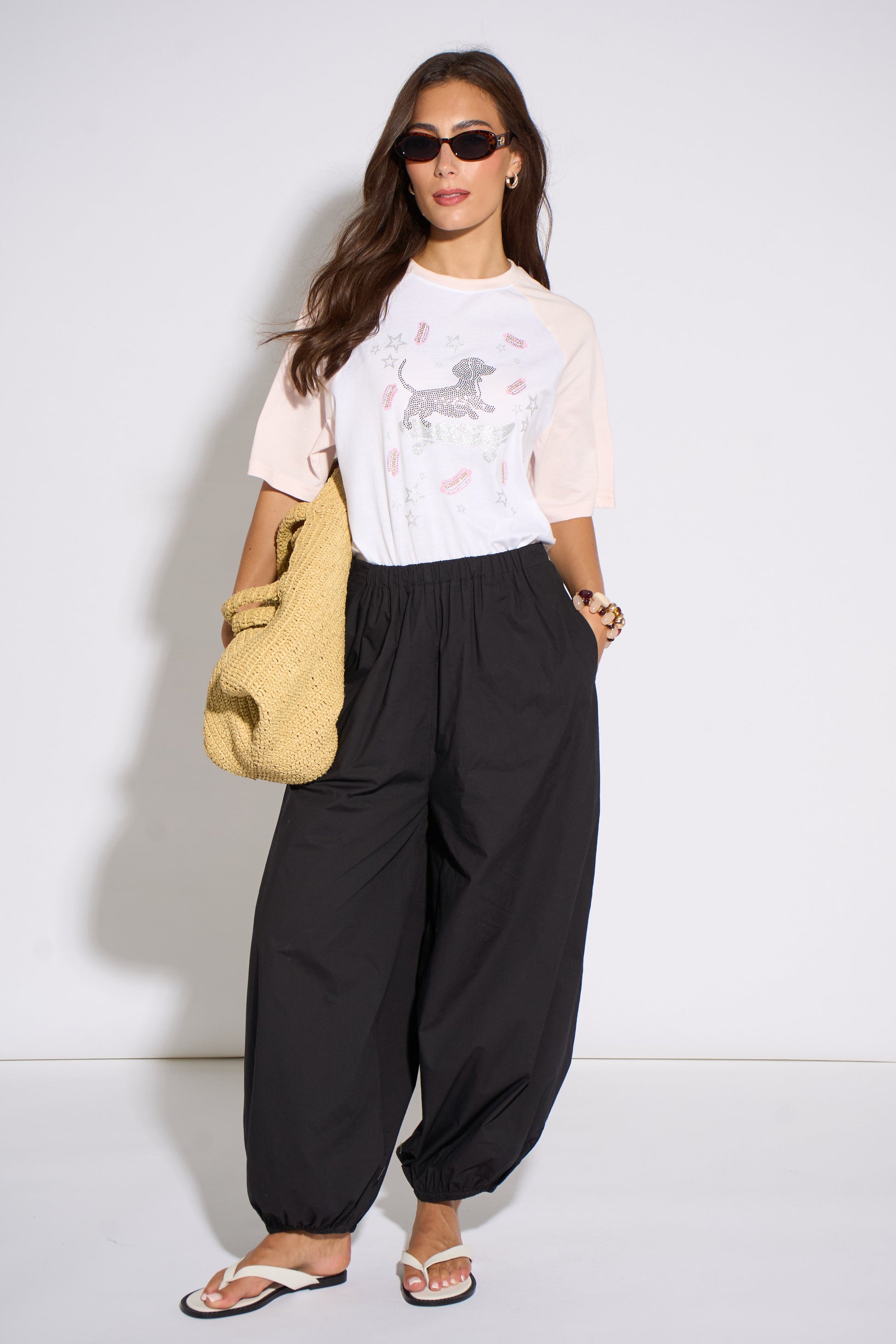 Woman wearing a white t-shirt with a graphic design and black pants, holding a yellow bag against a white background.