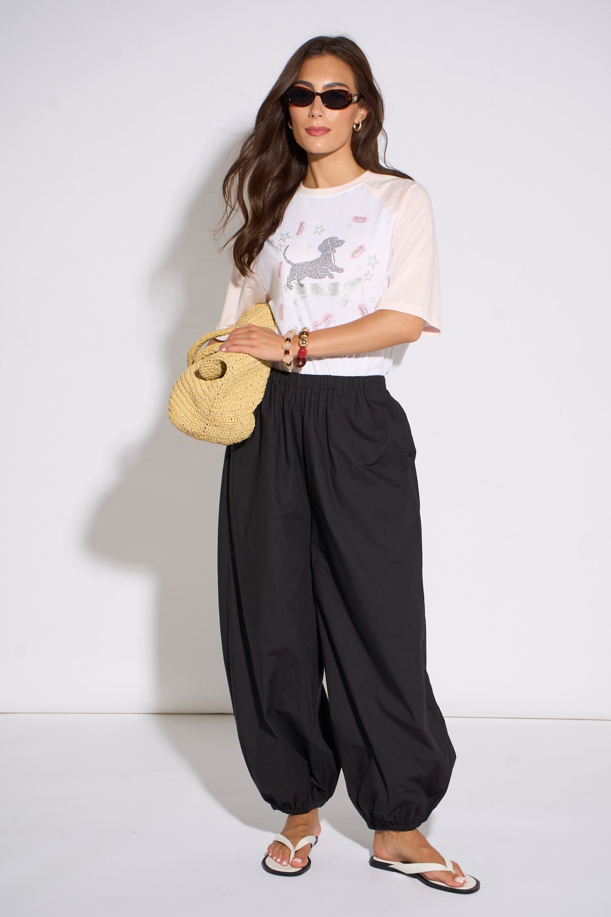Woman wearing a white top with a graphic design and black wide-leg pants, holding a straw bag against a white background.