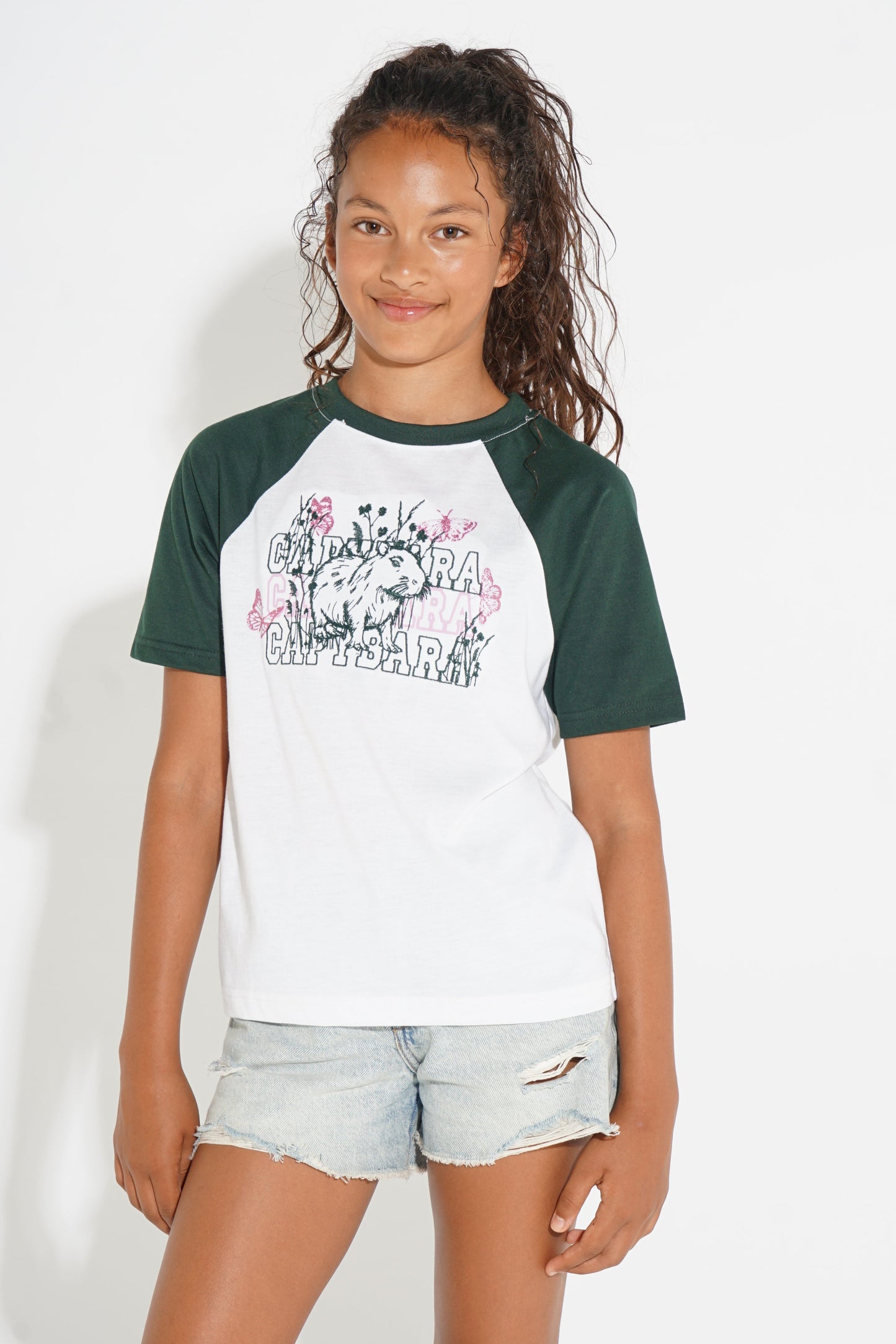 A young girl modeling a short-sleeve raglan baseball t-shirt with a white body, dark green sleeves, and a graphic print on the chest, paired with light-wash denim shorts against a plain white background.