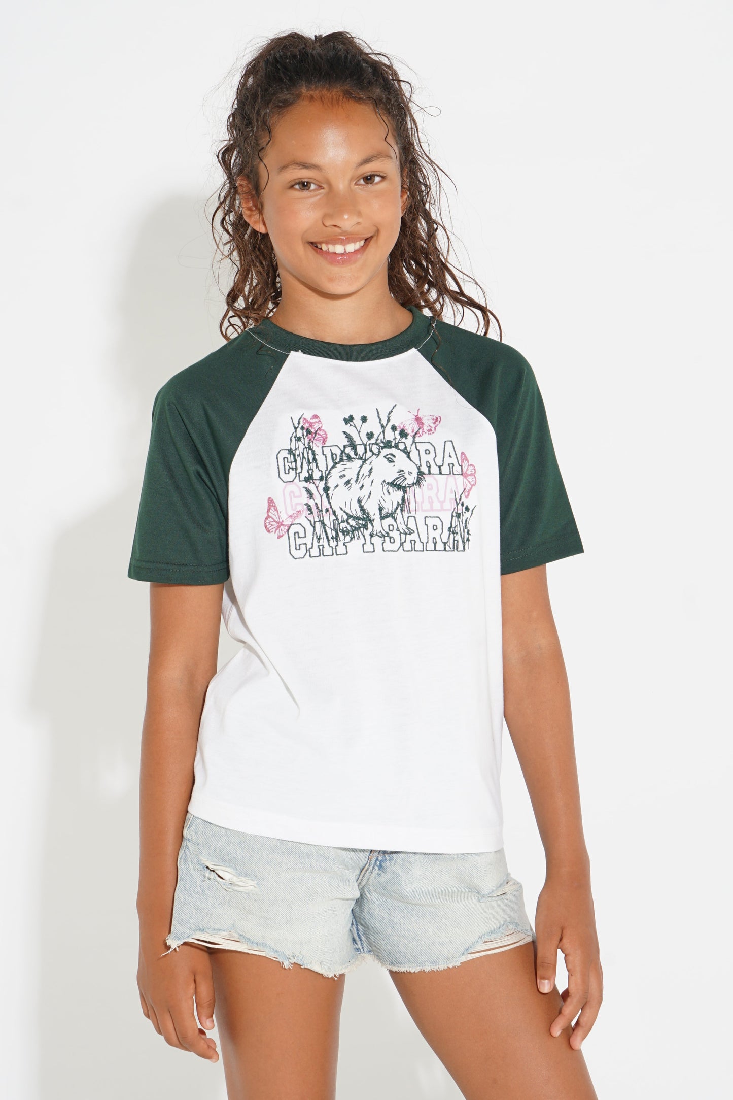 A young girl smiling and wearing a raglan-style baseball t-shirt with dark green sleeves and collar, and a white body featuring a graphic print of a lion and the words 'CANAAN CAN'T BE SEEN'. She is also wearing light-wash denim shorts against a plain white background.