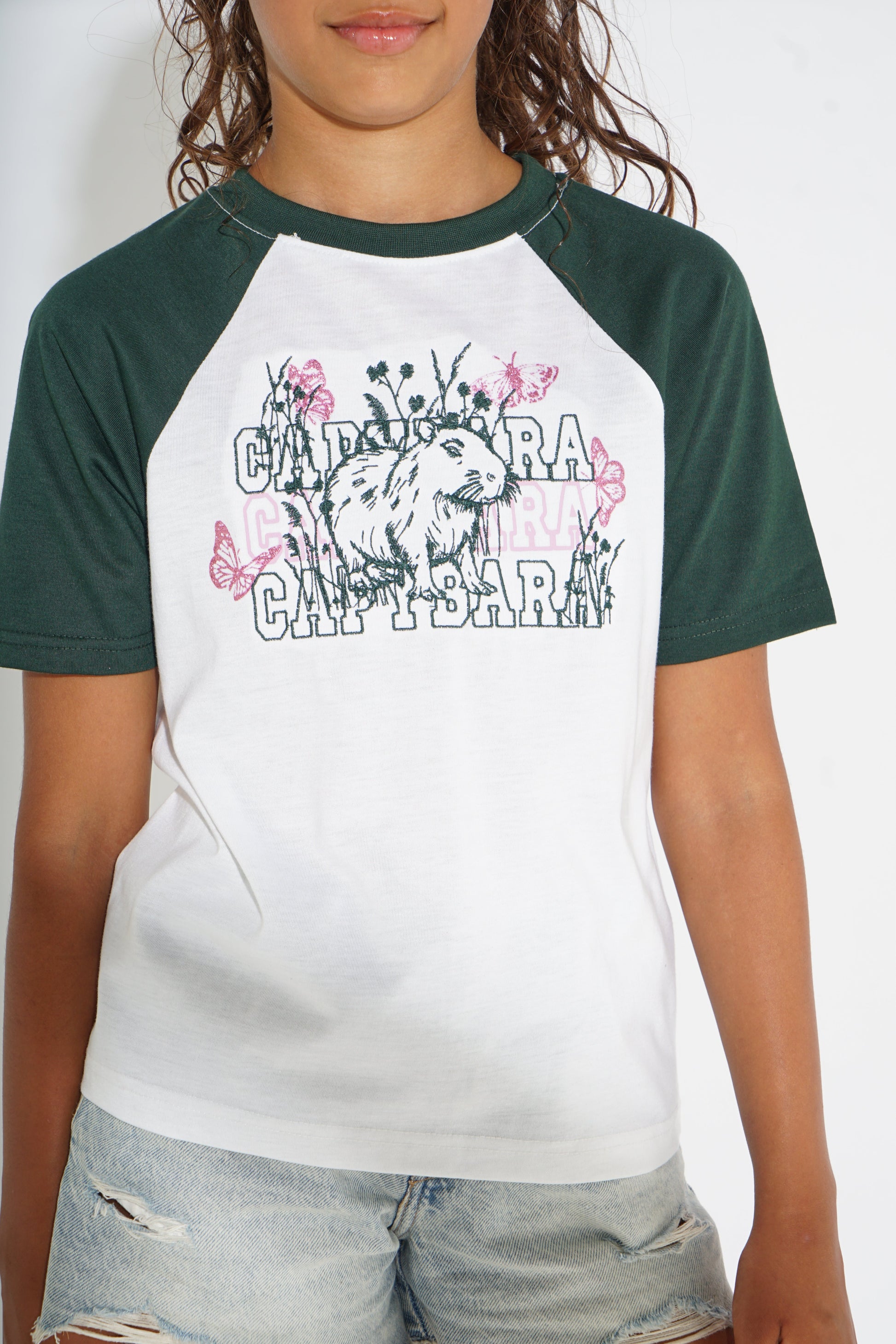 A child wearing a raglan baseball t-shirt with a white body and dark green sleeves, featuring a graphic print of a lion cub and the text 'CABITRA' and 'CABITRA GIRL T-SHIRT'.