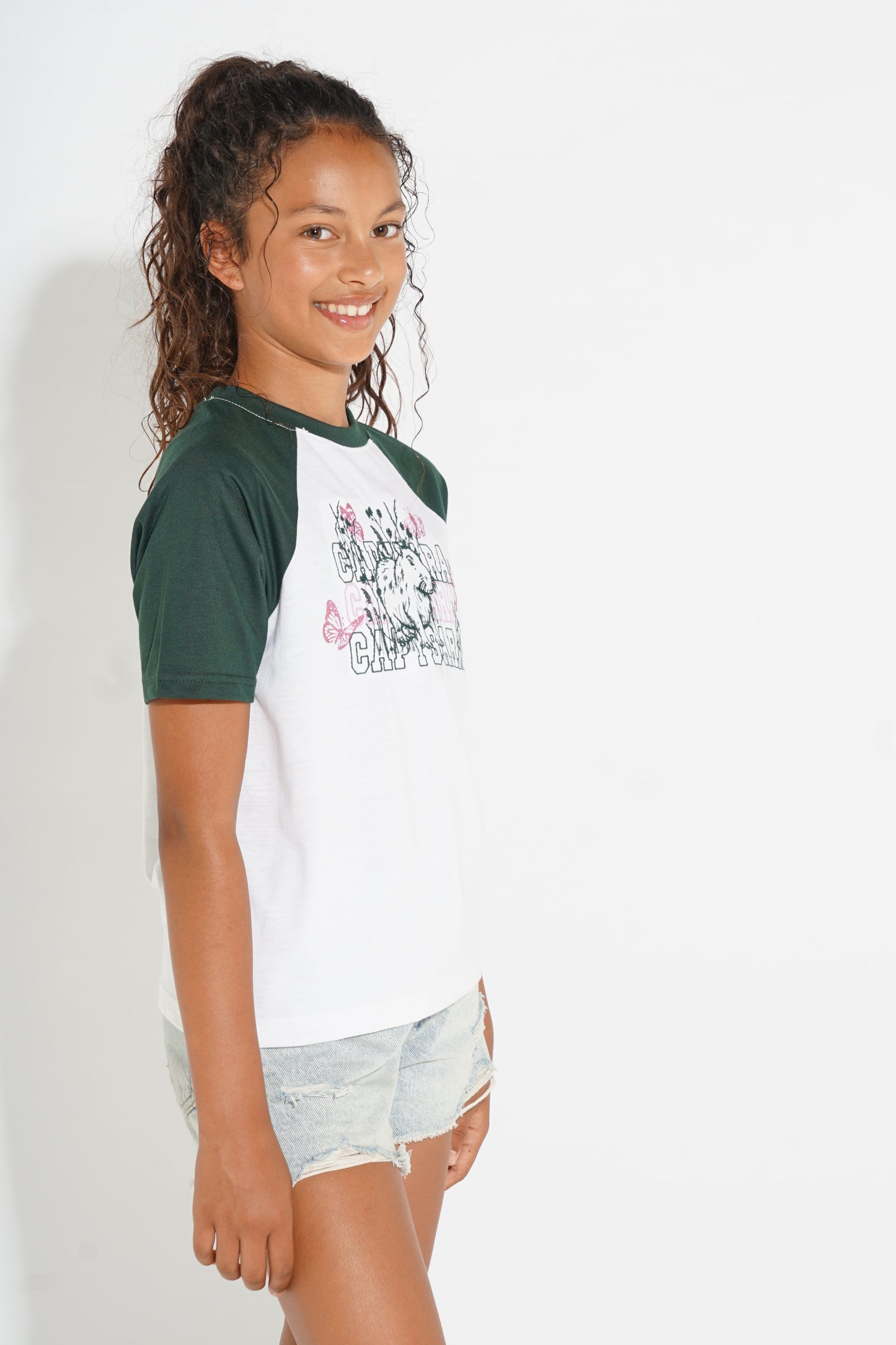 A young girl with curly brown hair tied back is shown from the waist up, wearing a raglan-style baseball t-shirt with dark green sleeves and a white body featuring a graphic print, paired with light-wash denim shorts, against a plain white background.