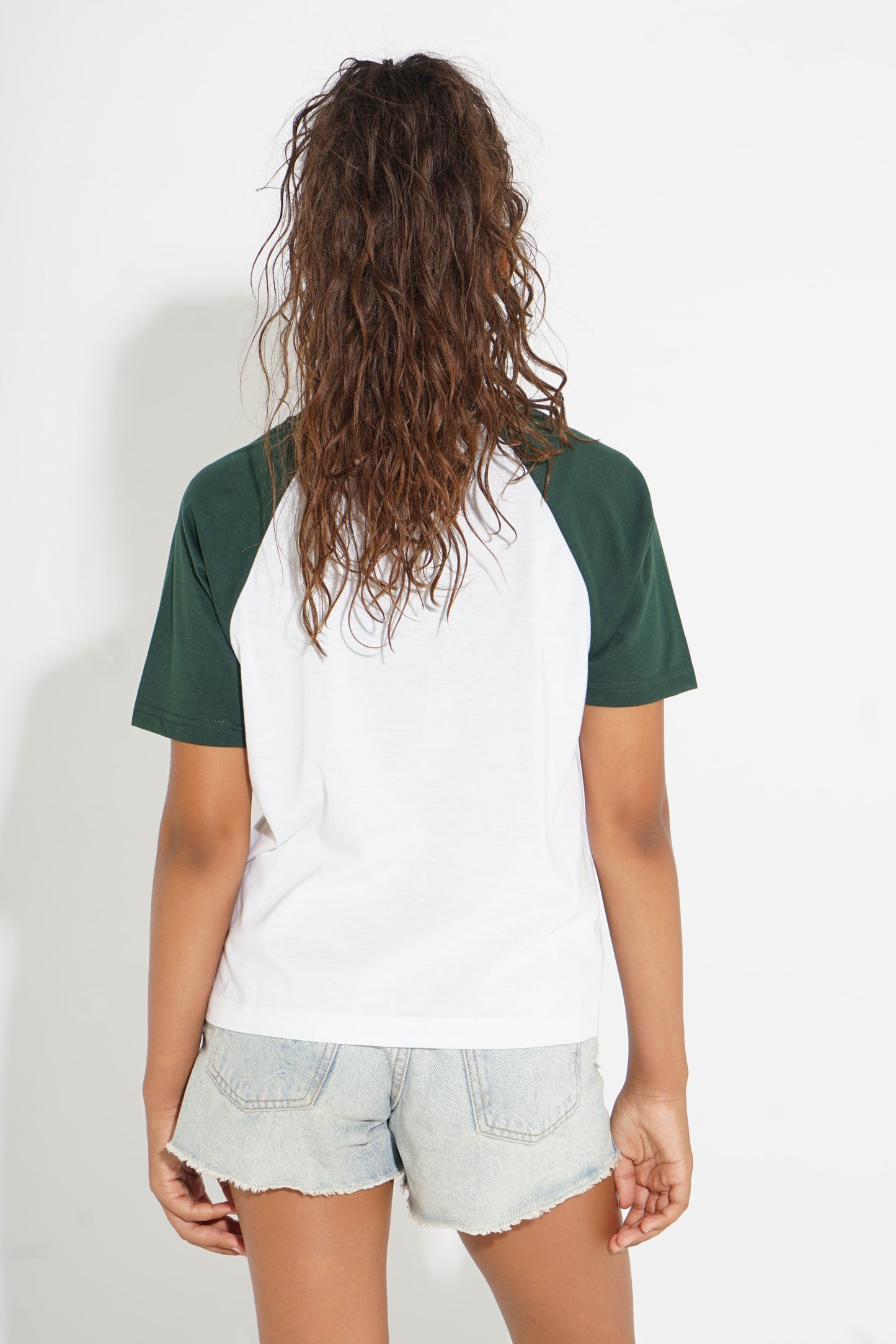 A person shown from the back wearing a raglan-style short-sleeve t-shirt with a white body and dark green sleeves, paired with light denim shorts against a plain white background.