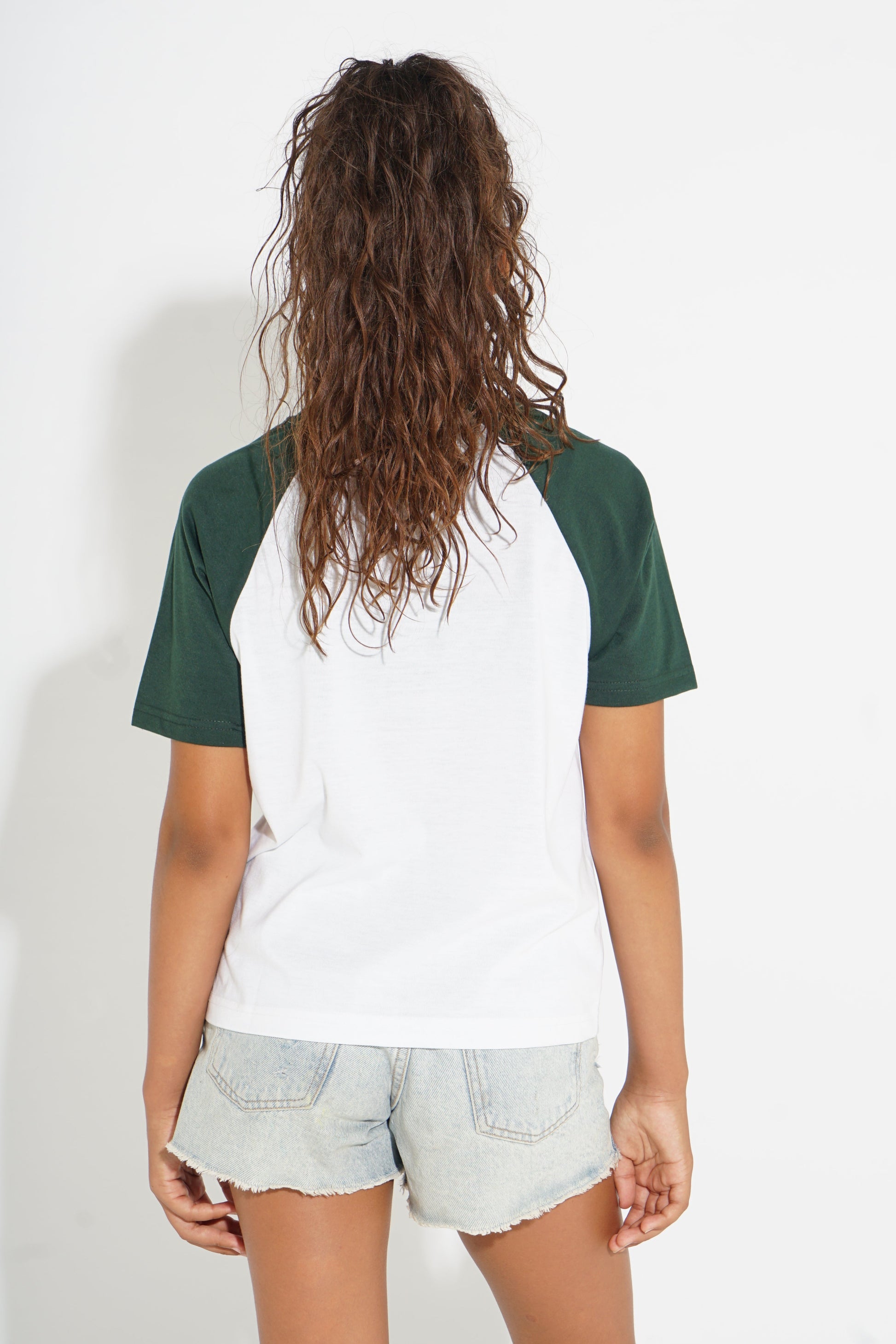 A person shown from the back wearing a raglan-style short-sleeve t-shirt with a white body and dark green sleeves, paired with light denim shorts against a plain white background.