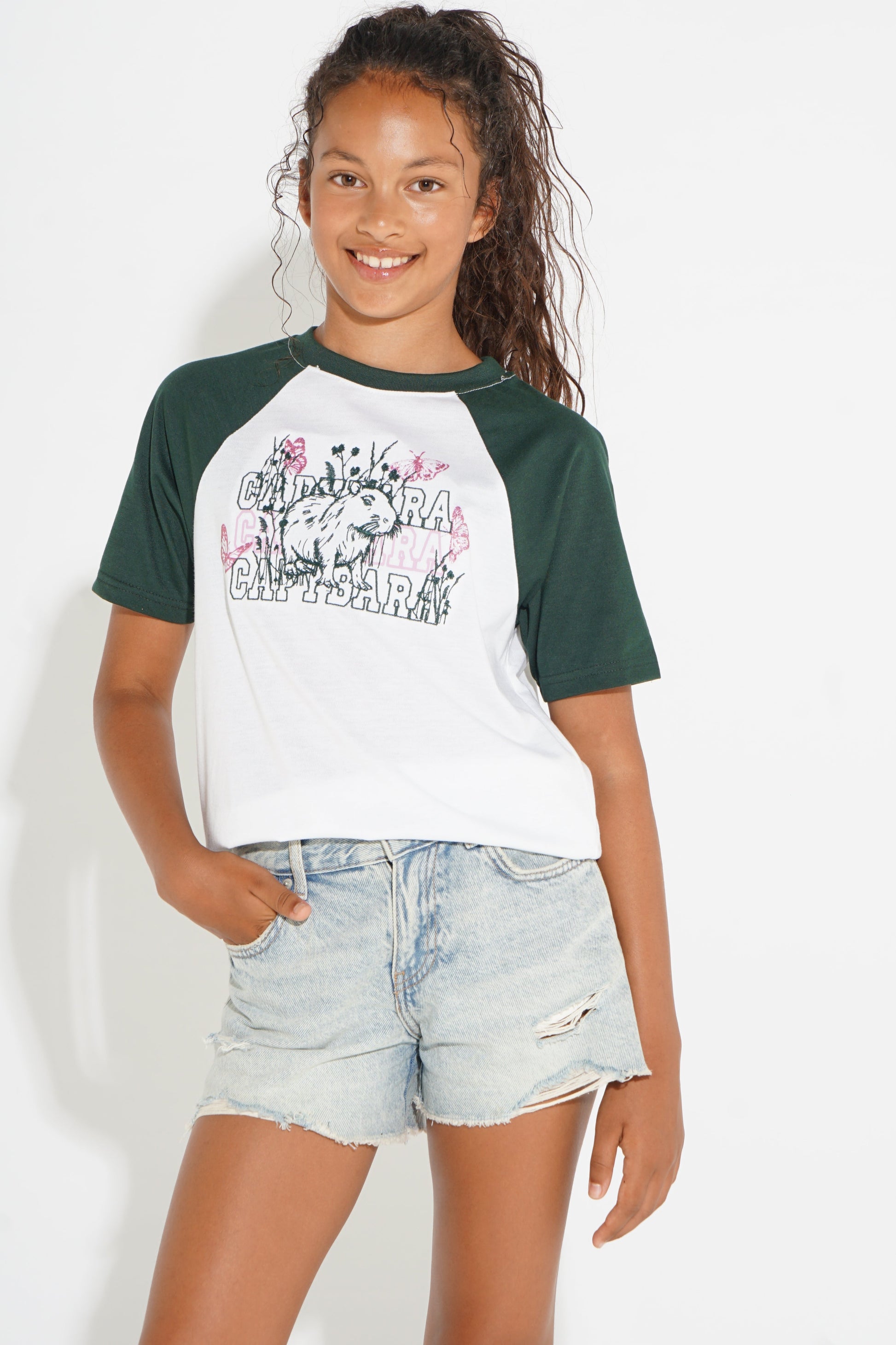 A young girl modeling a short-sleeve raglan t-shirt with dark green sleeves and a white body featuring a colorful graphic print, paired with light-wash denim shorts against a plain white background.