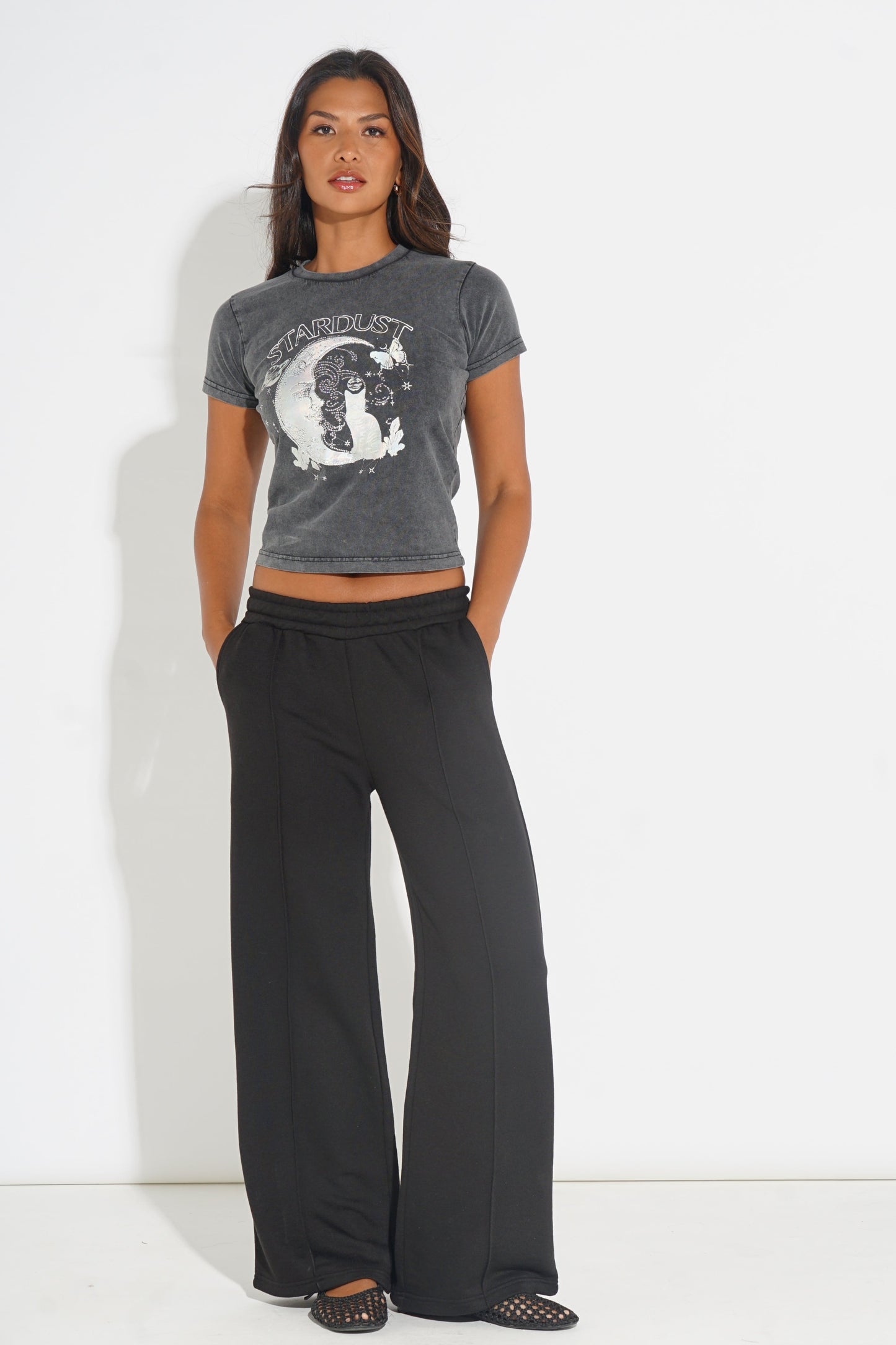 A woman modeling a dark gray cropped t-shirt with a white moon graphic and the word 'FANTASTIC' on the front, paired with black wide-leg sweatpants and black flat shoes, standing against a white studio background.