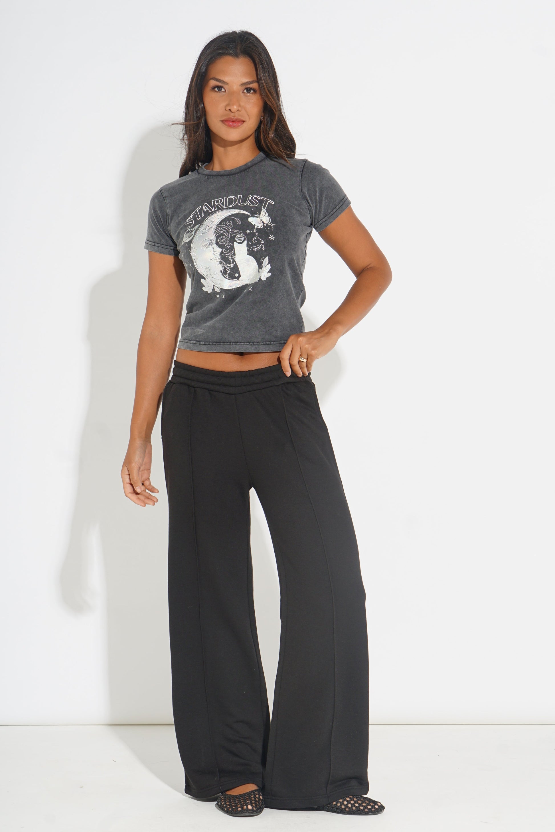 A woman modeling a dark gray graphic crop top and black wide-leg pants against a plain white studio background.