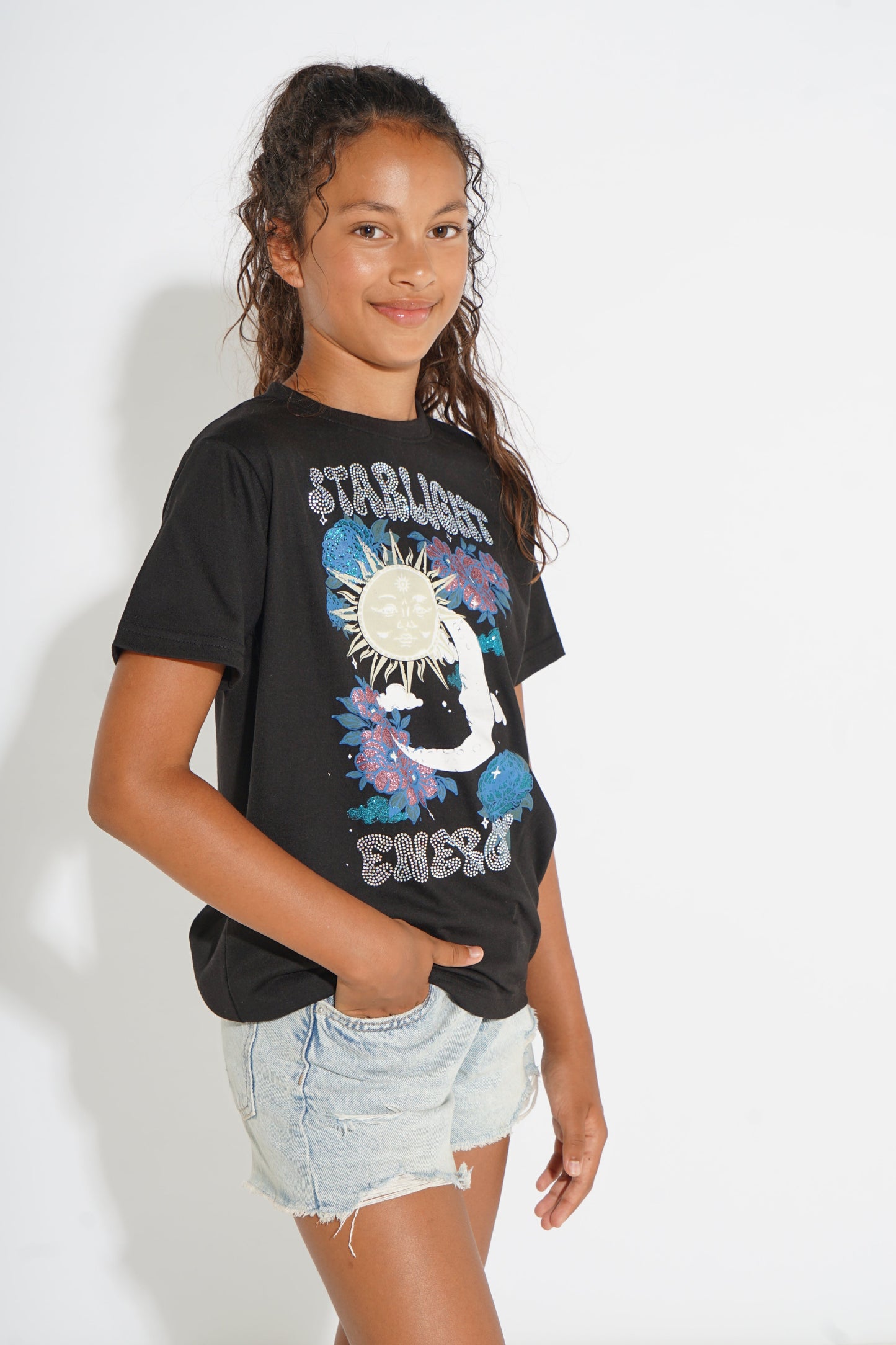 A girl modeling a black graphic t-shirt with a colorful sun and moon design and the words 'STARLIGHT' and 'ENERGY', paired with light-wash denim shorts against a white studio background.