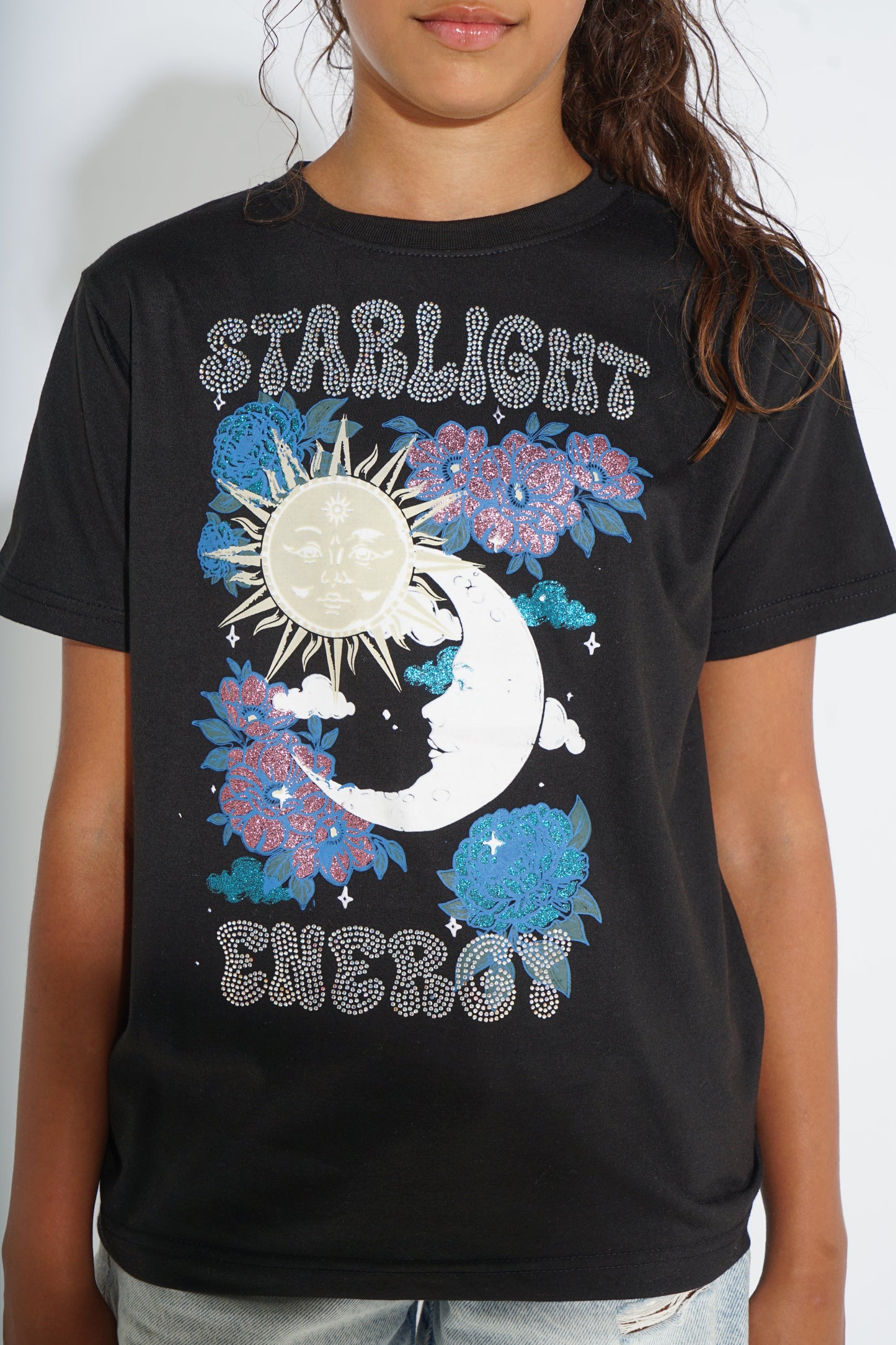 A black short-sleeve graphic t-shirt featuring a celestial design with a sun, crescent moon, and blue flowers, with the text 'STARLIGHT ENERGY' in a rhinestone-like pattern. The shirt is modeled by a person against a plain light background.