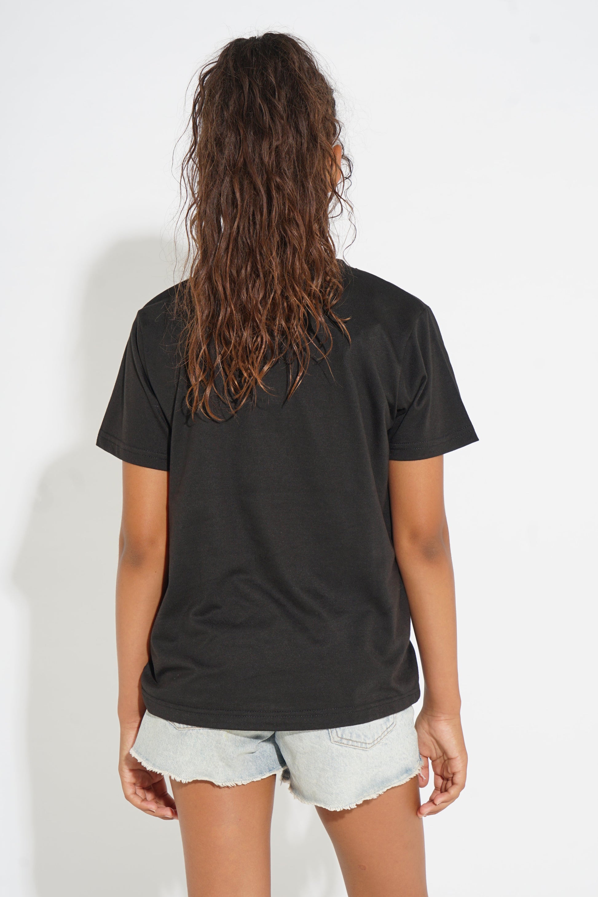 A woman viewed from behind wearing a black short-sleeve t-shirt and light-wash denim shorts against a plain white studio background.