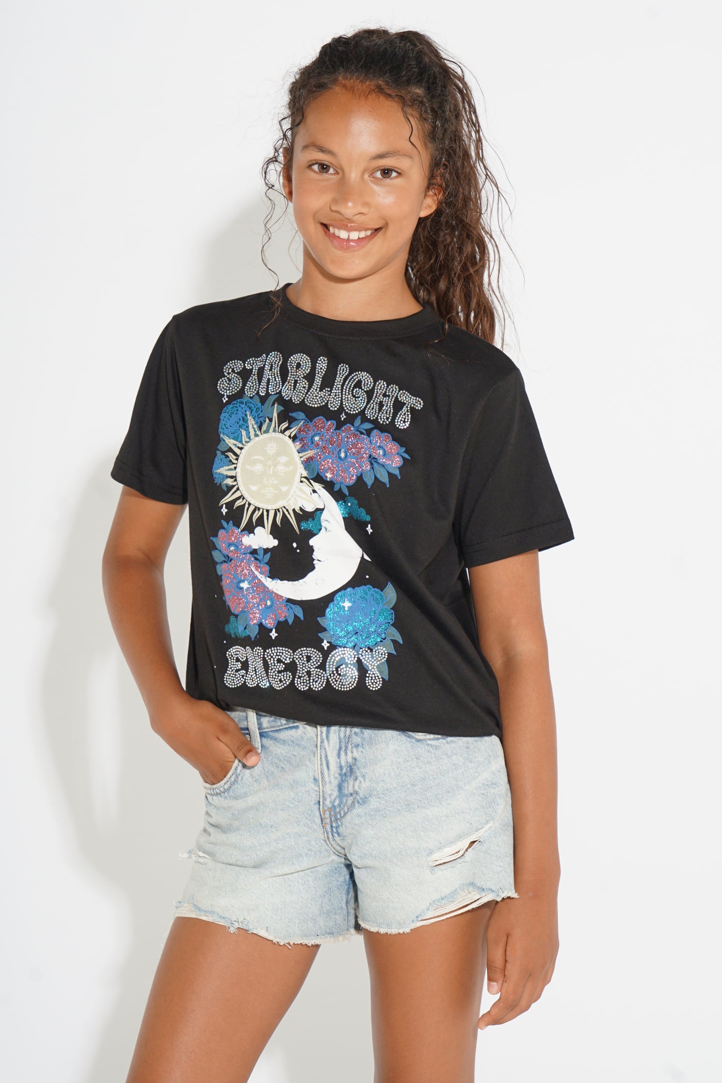 A young girl with dark curly hair smiling, wearing a black graphic t-shirt with 'STIRLIGHT ENERGY' and a moon/sun design, paired with light-wash denim shorts, against a plain white background.