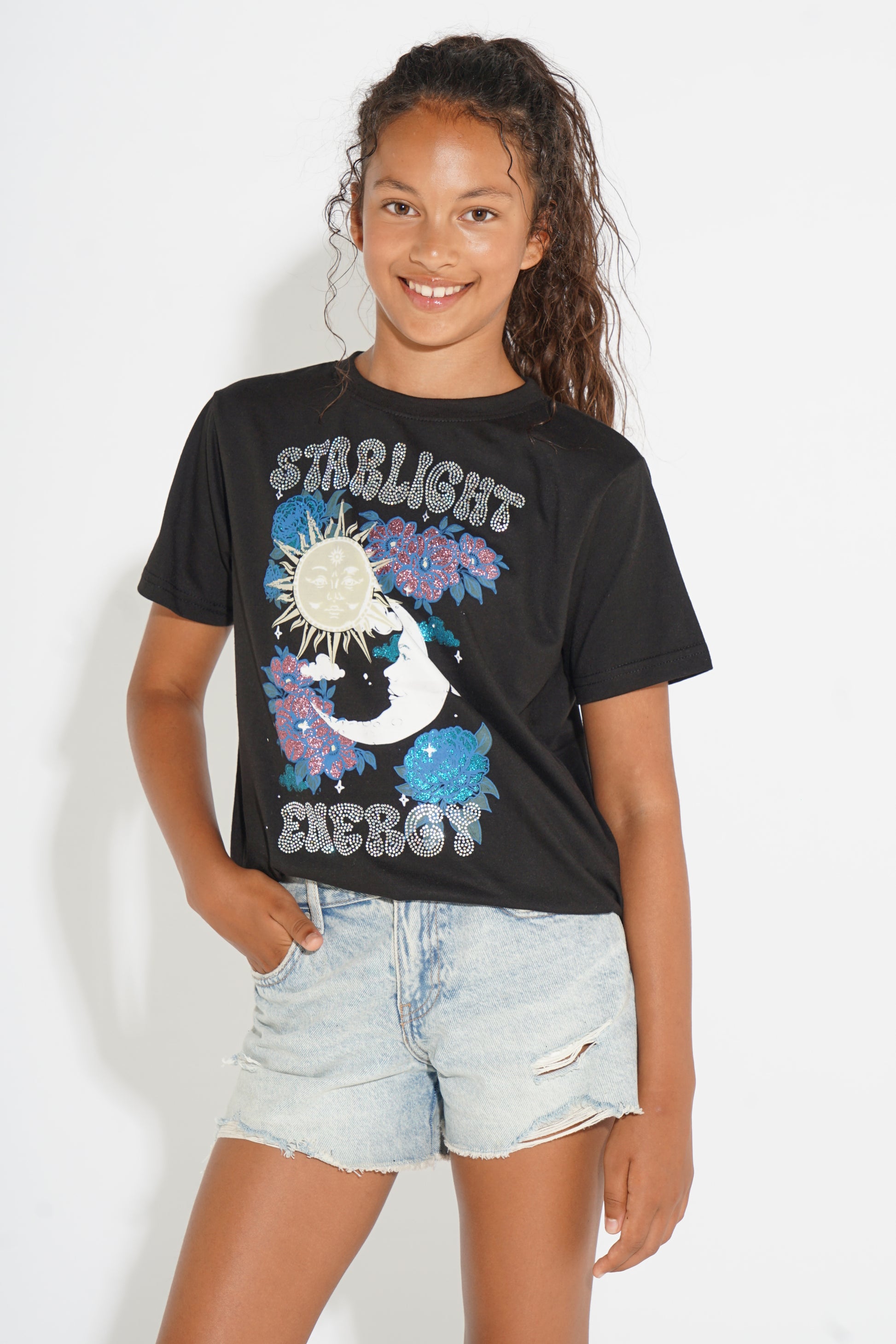 A young girl with dark curly hair smiling, wearing a black graphic t-shirt with 'STIRLIGHT ENERGY' and a moon/sun design, paired with light-wash denim shorts, against a plain white background.