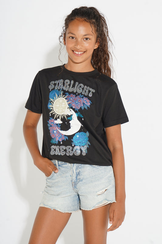 A young girl with dark curly hair smiling, wearing a black graphic t-shirt with 'STIRLIGHT ENERGY' and a moon/sun design, paired with light-wash denim shorts, against a plain white background.