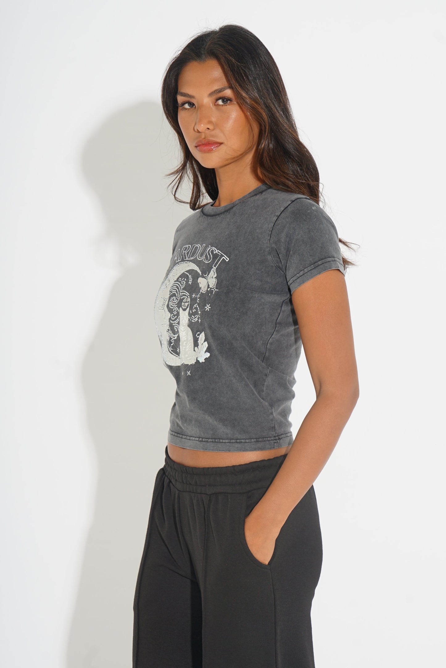 A woman modeling a dark gray acid-wash cropped t-shirt with a white graphic print, paired with black sweatpants, against a plain white studio background.