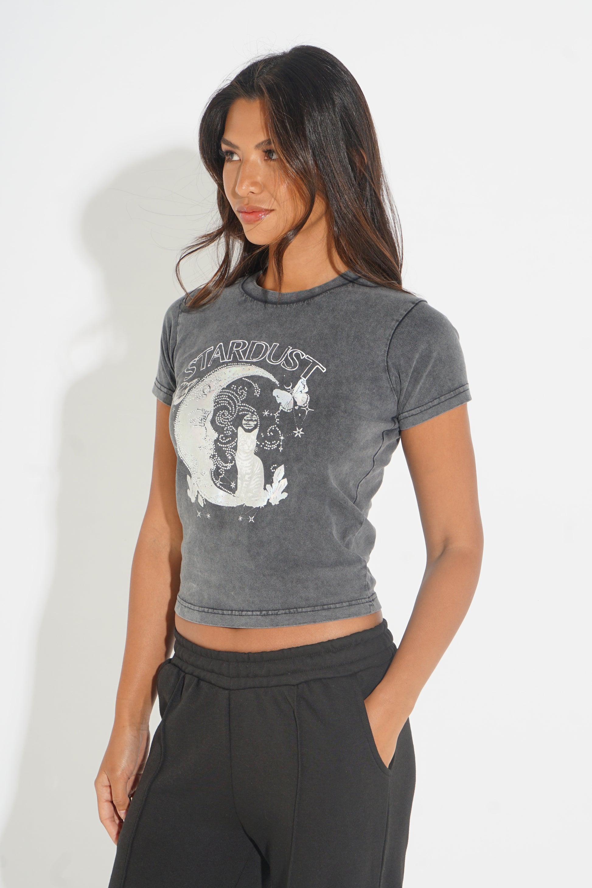 A woman modeling a dark gray acid-wash cropped t-shirt with a white 'Stardust' cat graphic, paired with black sweatpants against a white studio background.