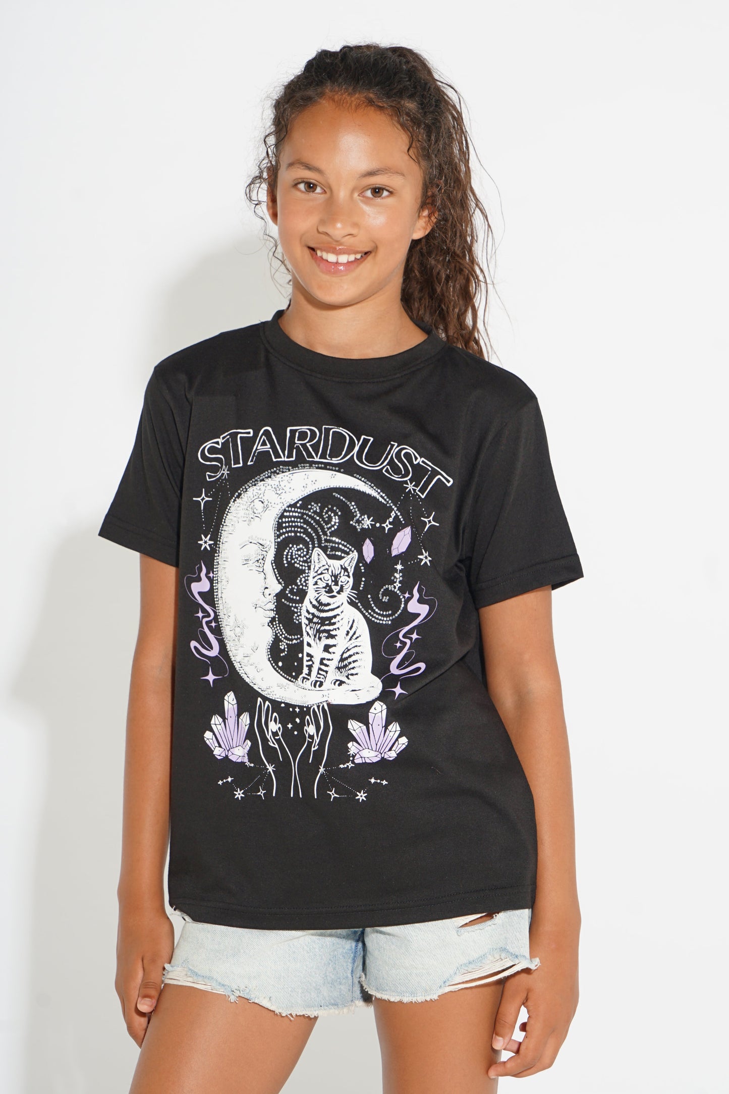 A young girl with curly brown hair wearing a black graphic t-shirt with a white and purple celestial design that reads 'STARDUST', paired with light-wash denim shorts, standing against a plain white background.