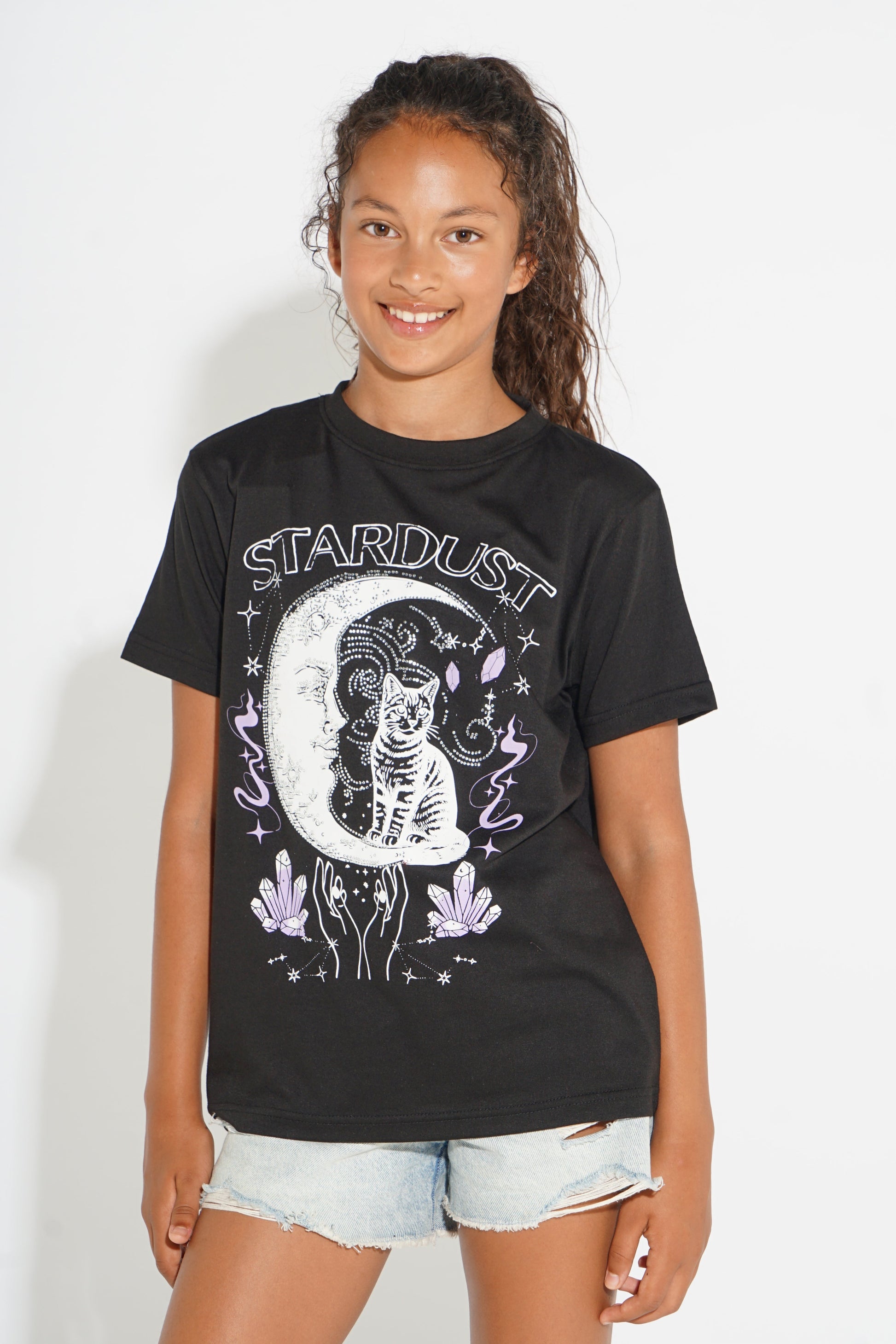 A young girl with curly brown hair wearing a black graphic t-shirt with a white and purple celestial design that reads 'STARDUST', paired with light-wash denim shorts, standing against a plain white background.