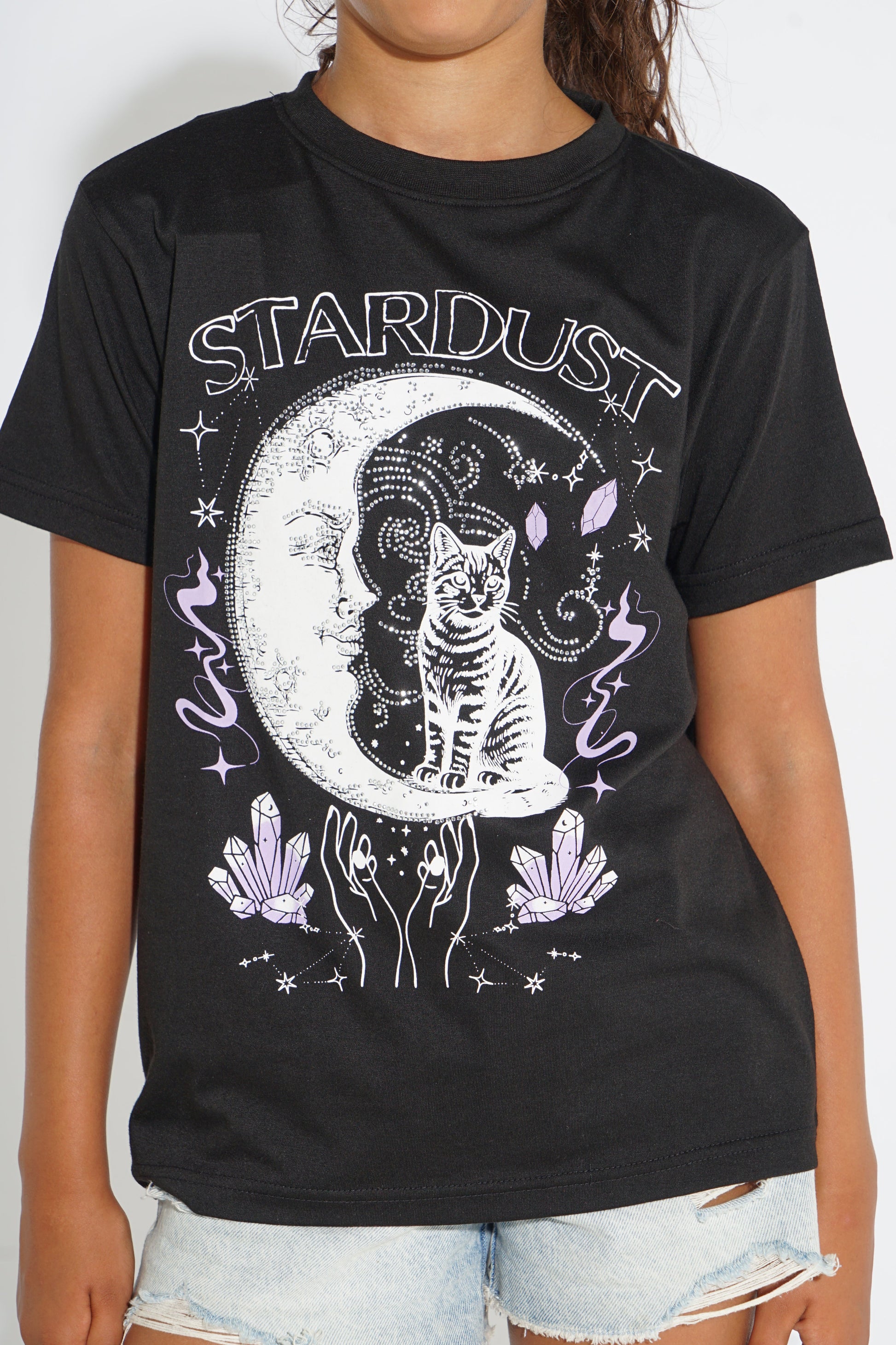 A person wearing a black graphic T-shirt with a white and lavender celestial print featuring a cat, crescent moon, crystals, and the word 'STARDUST', paired with light denim shorts.