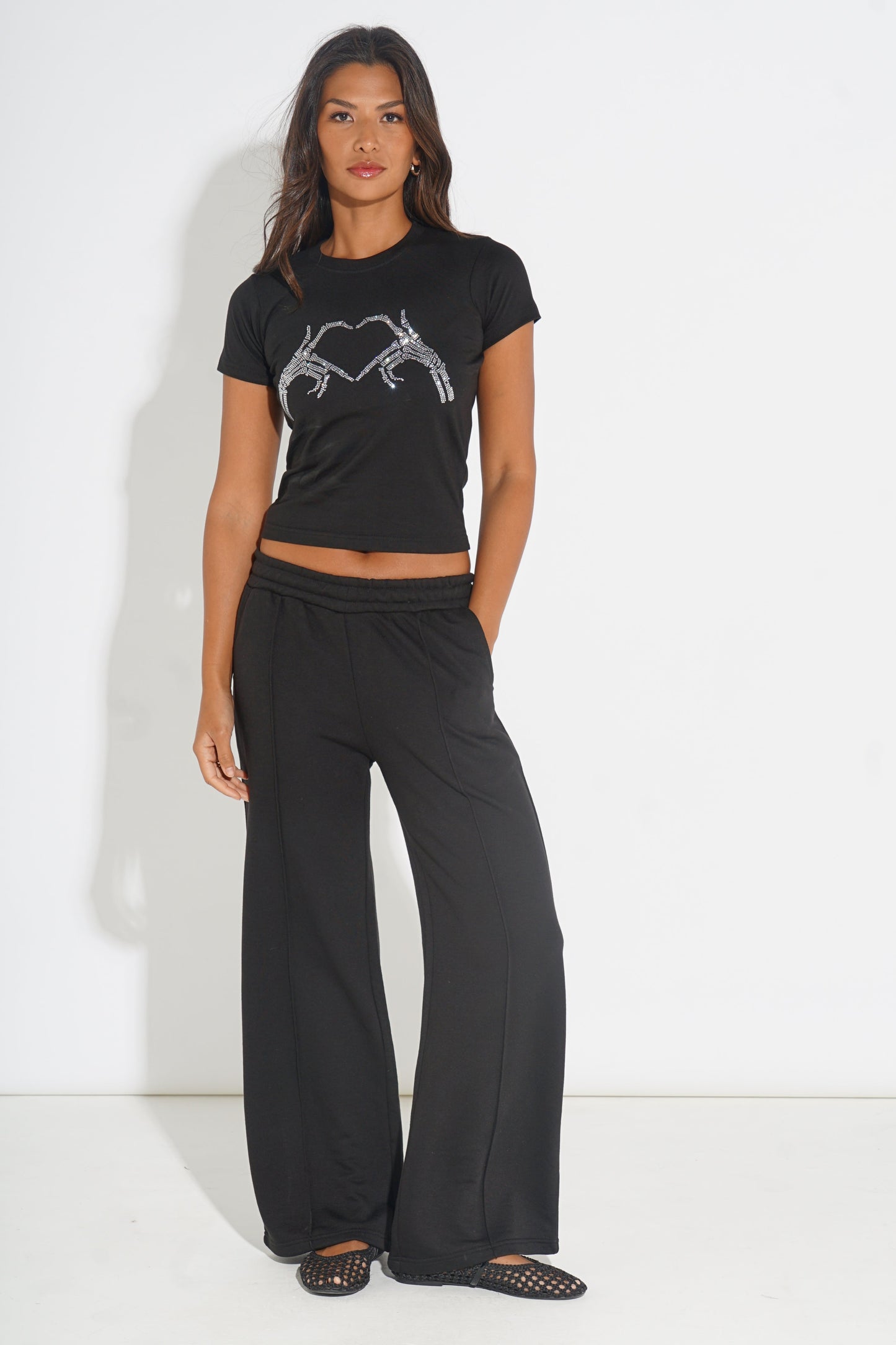 A woman modeling a black short-sleeve crop top with a silver rhinestone heart design on the chest, paired with black wide-leg pants and black open-toe flat shoes, set against a plain white studio background.