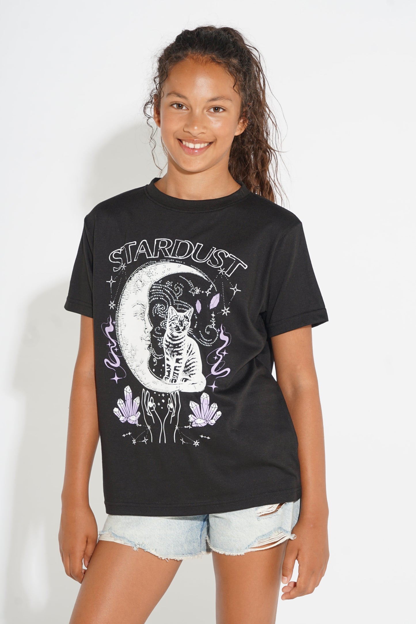 A young girl smiling and wearing a black graphic t-shirt with a white skeleton cat and moon design, paired with light-wash denim shorts, against a plain white background.