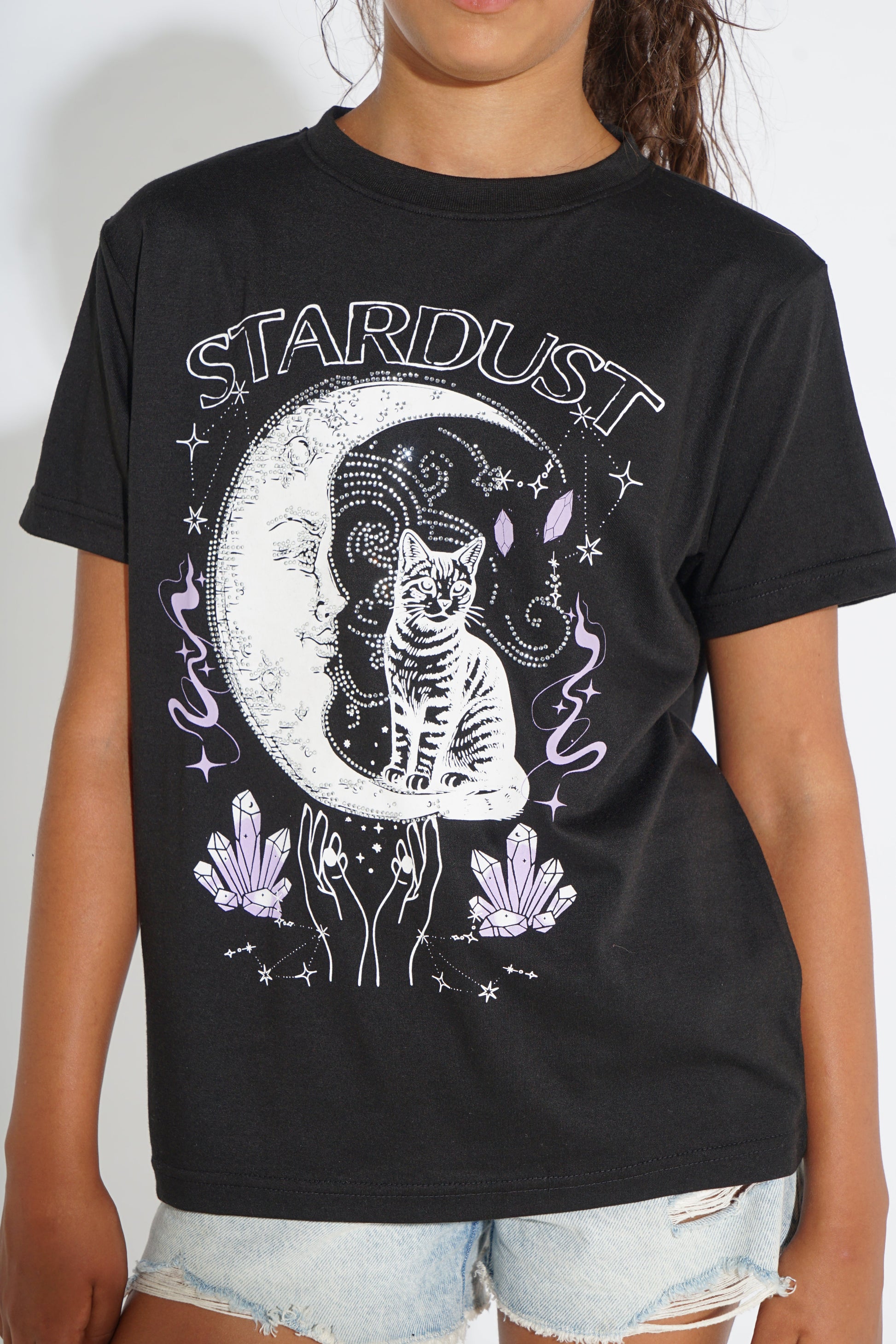A person wearing a black graphic T-shirt with a white and lavender celestial cat design and the word 'STARDUST' across the chest, paired with light denim shorts against a plain light background.