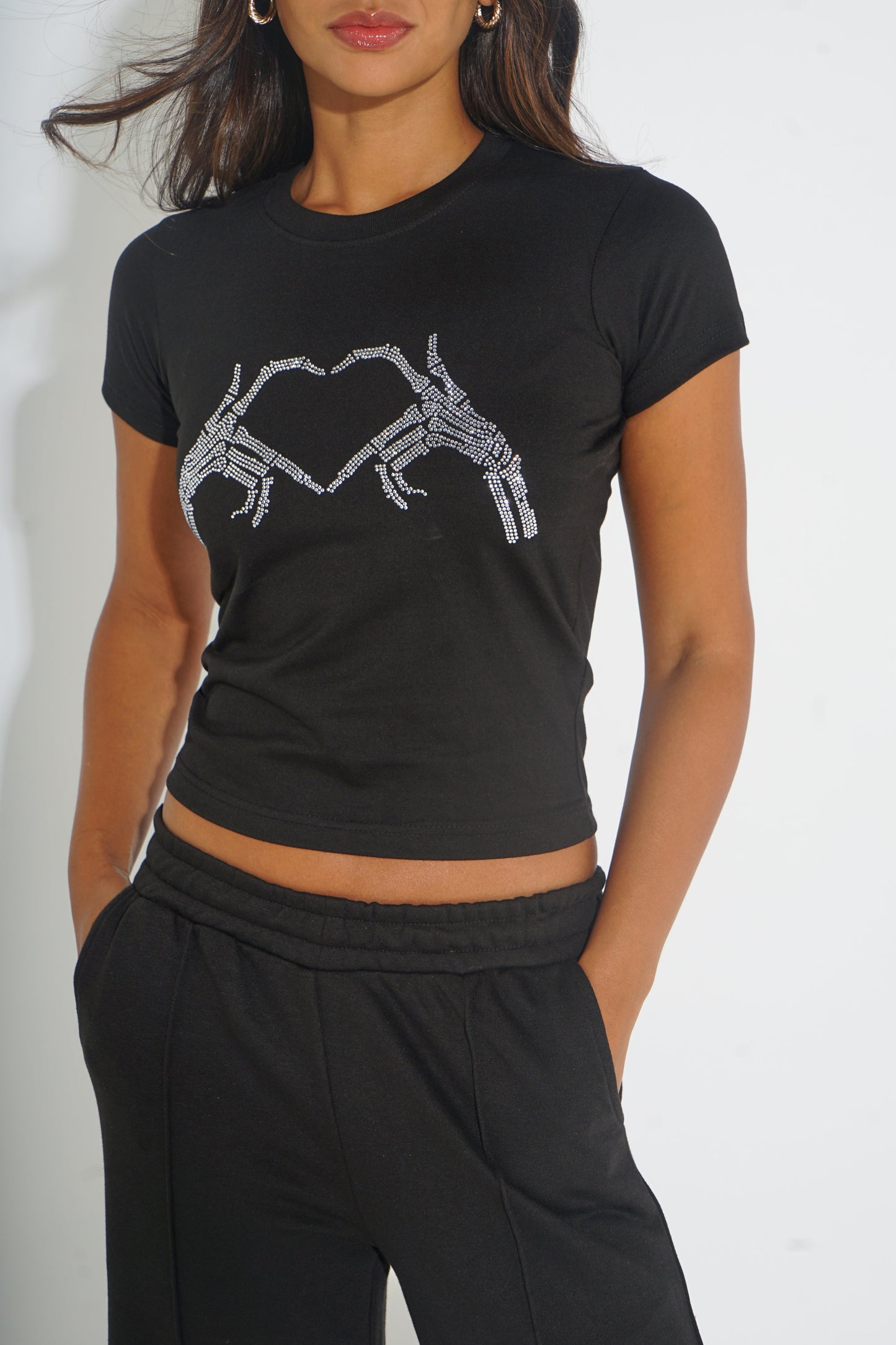 A woman wearing a black short-sleeve crop top with a white skeleton hand graphic on the front, paired with black sweatpants, against a plain white background.