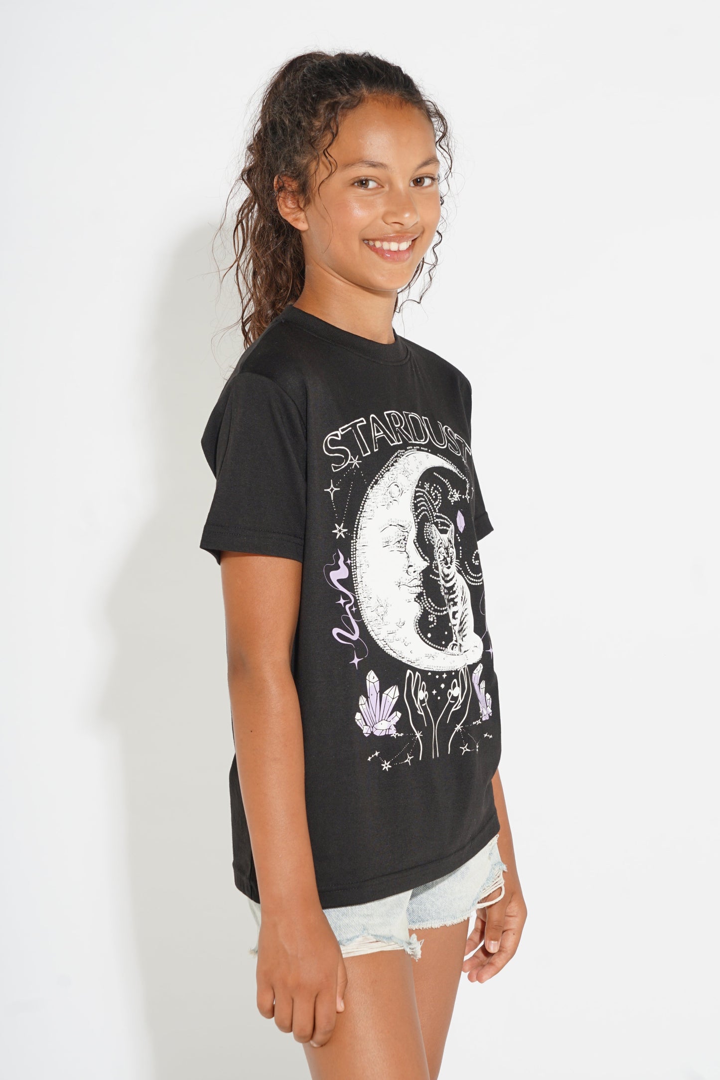 A young girl with her hair in a ponytail models a black graphic t-shirt featuring a white celestial design with the text 'STARBORN' and a skeleton illustration, paired with light denim shorts against a plain white background.