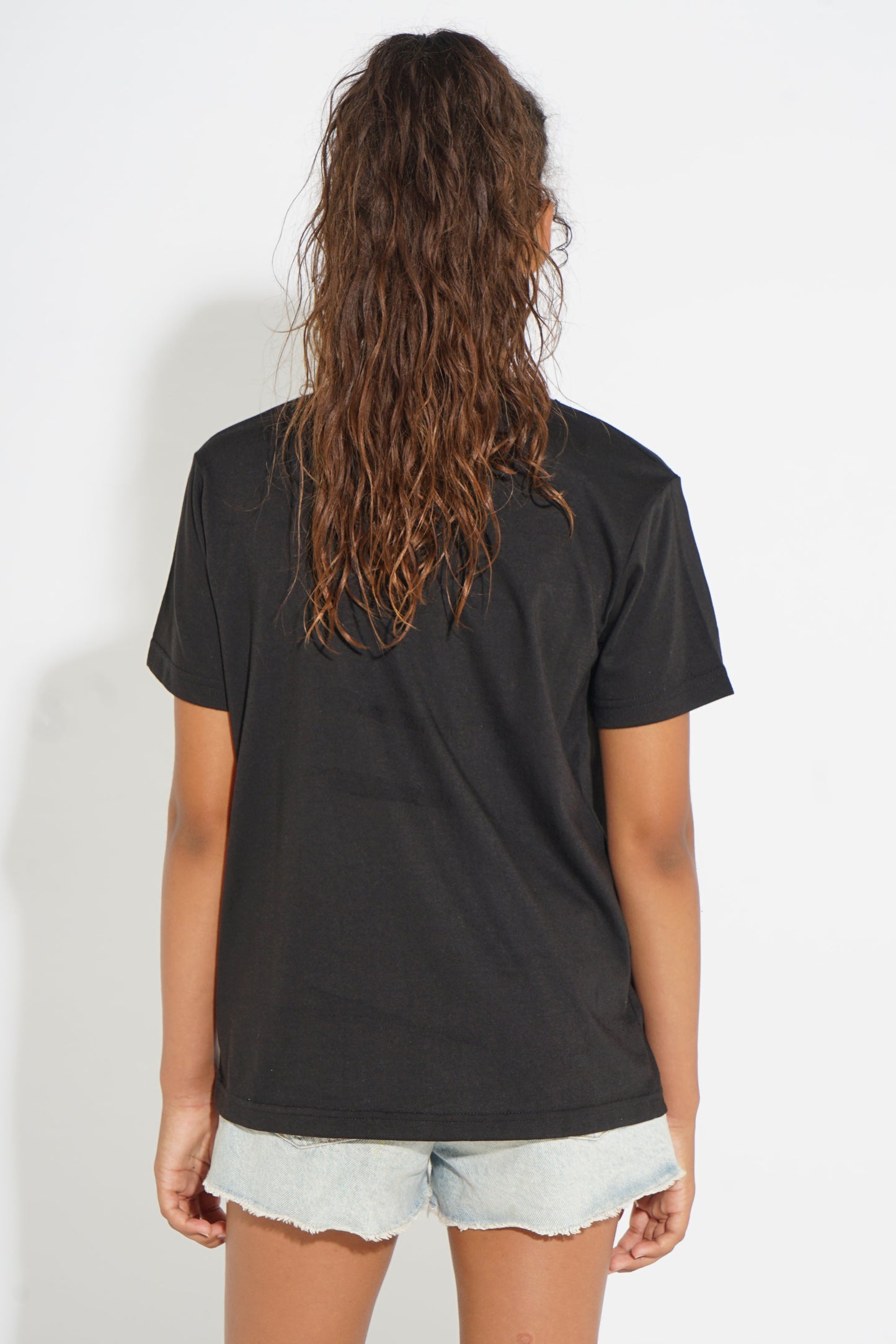 A woman viewed from behind wearing a black short-sleeve t-shirt and light-wash denim shorts against a plain white background.