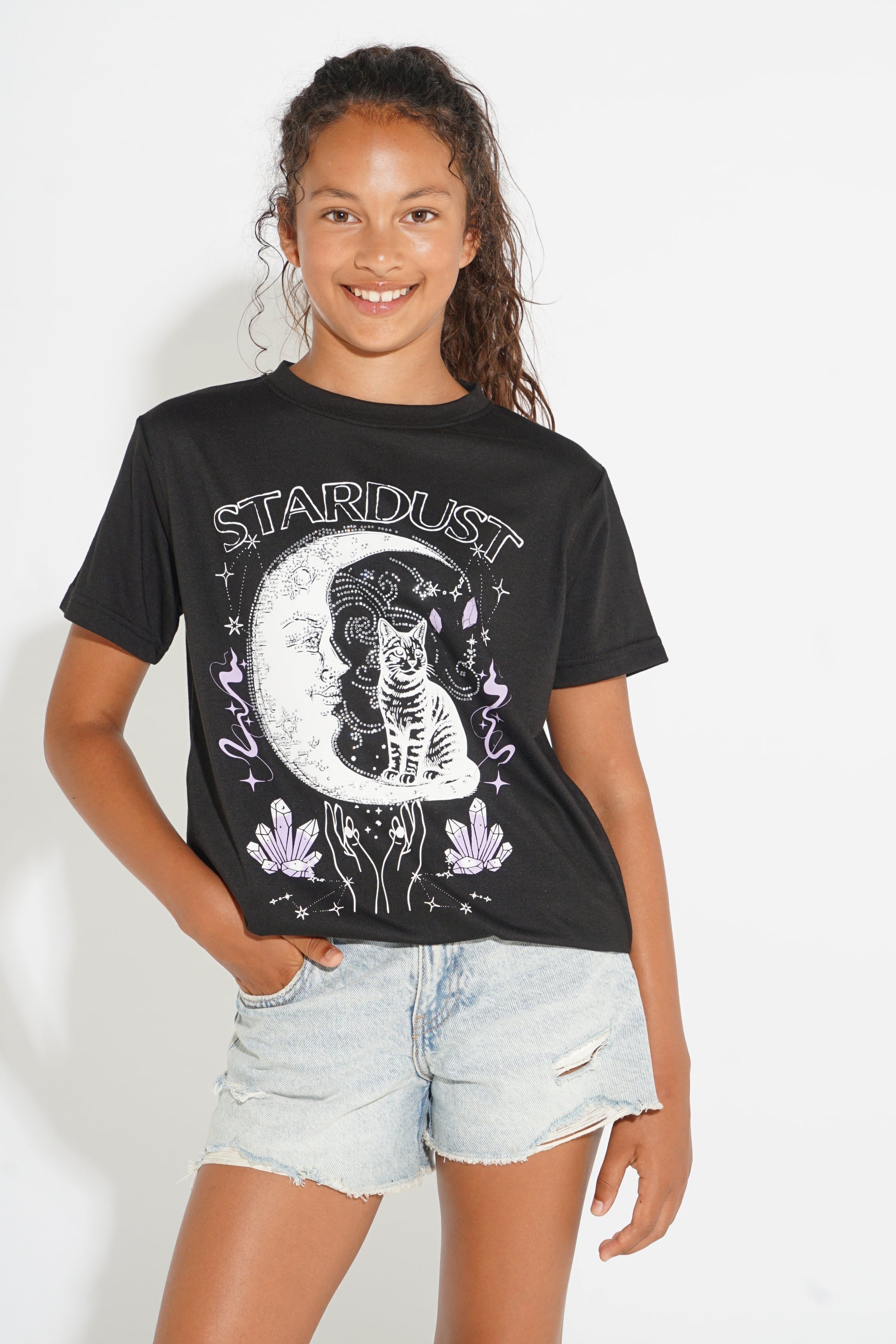 A young woman wearing a black graphic t-shirt with a white cat and moon design, paired with light-wash denim shorts, standing against a plain white background.