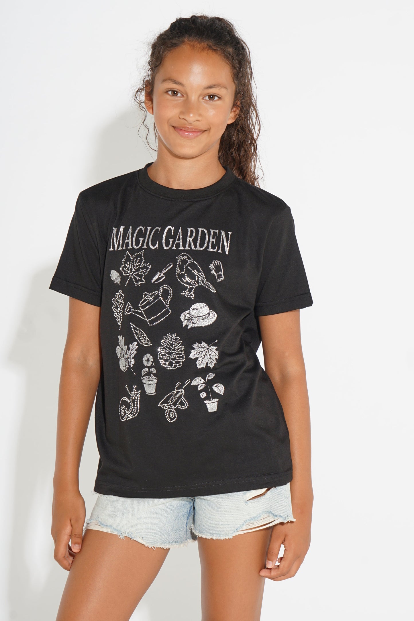 A young girl modeling a black short-sleeve graphic t-shirt with 'MAGIC GARDEN' printed in white across the chest, paired with light-wash denim shorts, against a plain white background.