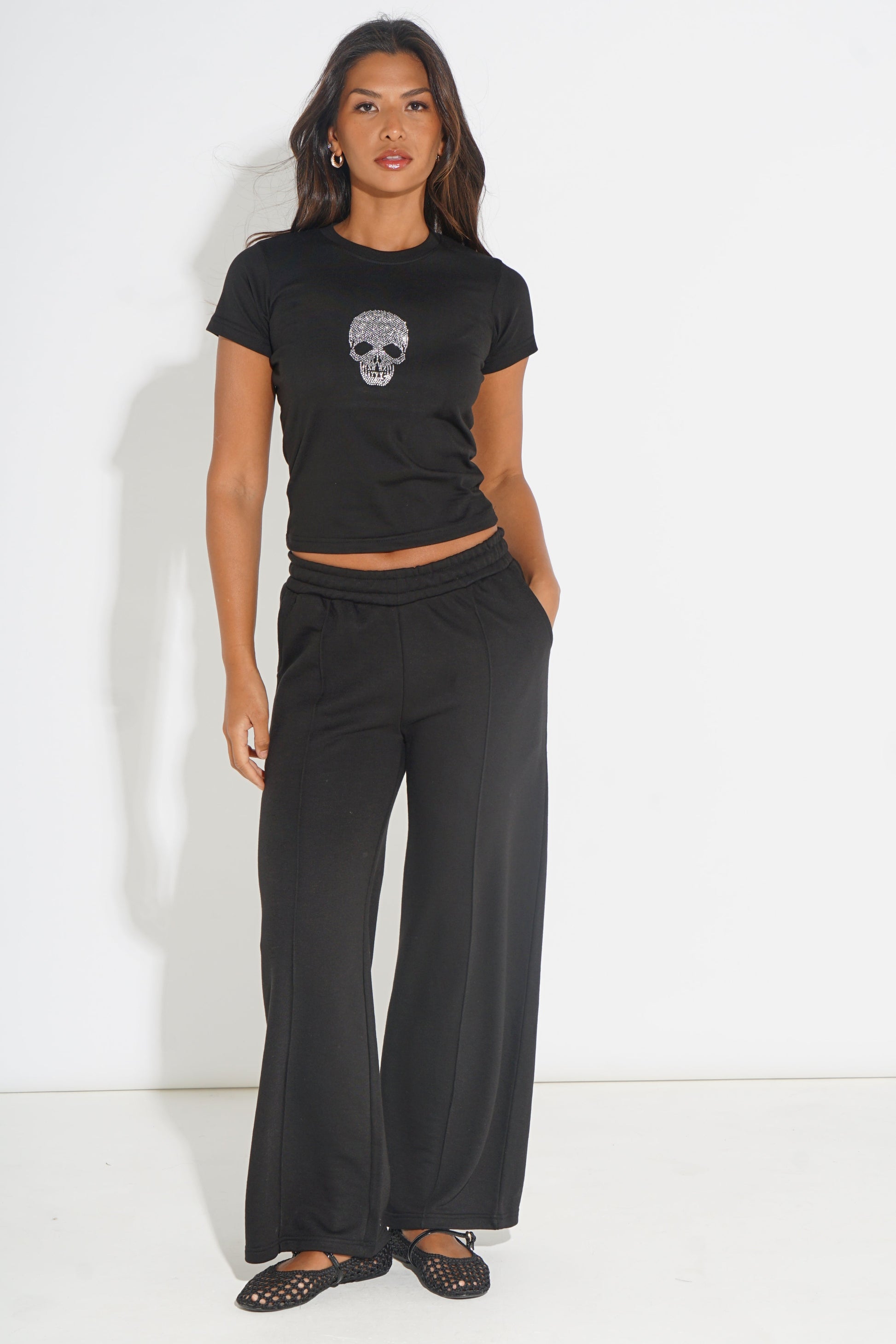 A woman modeling a black short-sleeve crop top with a skull graphic and matching black wide-leg pants against a plain white studio background.
