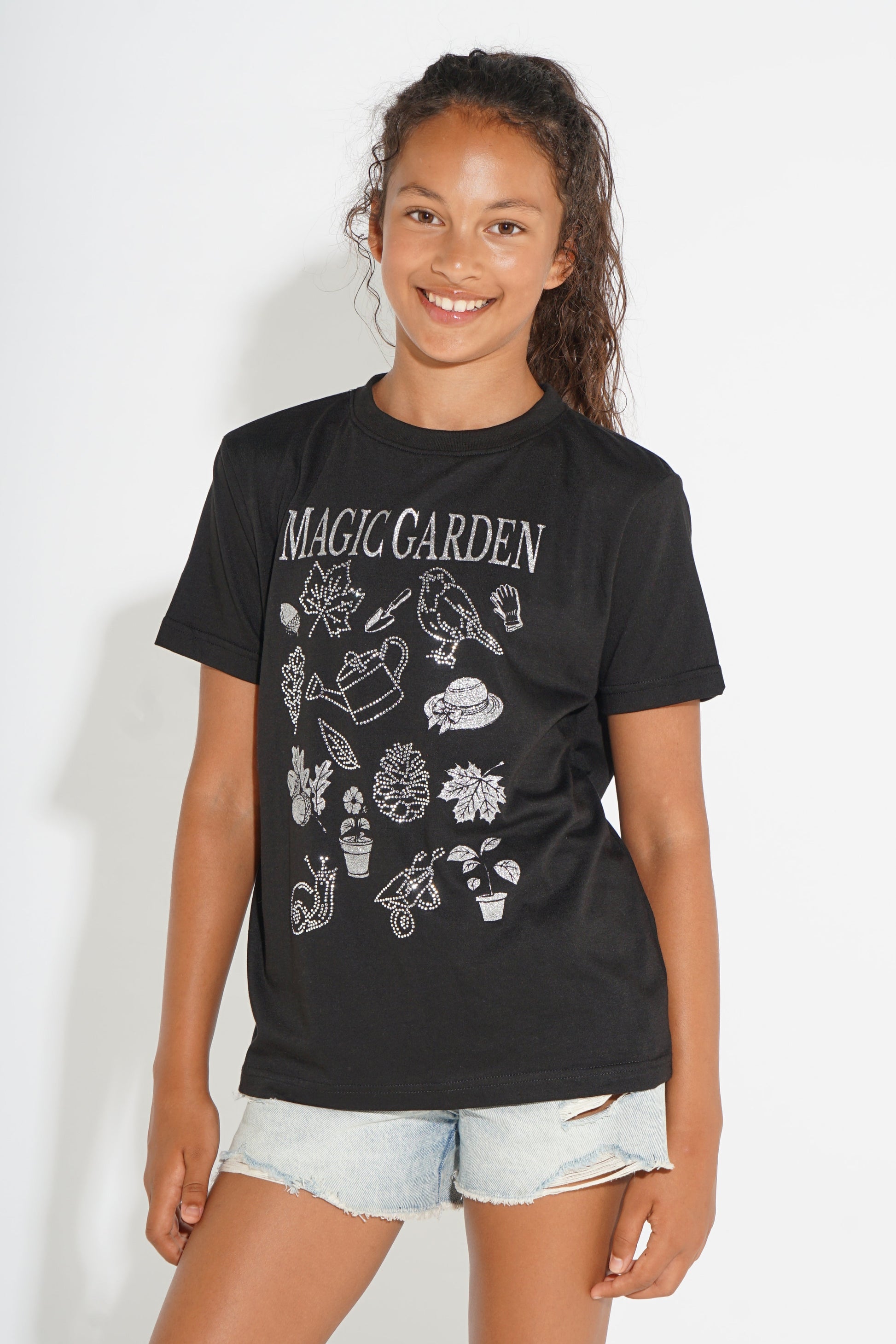 A young girl smiling and wearing a black graphic t-shirt with 'MAGIC GARDEN' printed in white text above gardening illustrations, paired with light-wash denim shorts, against a plain white background.