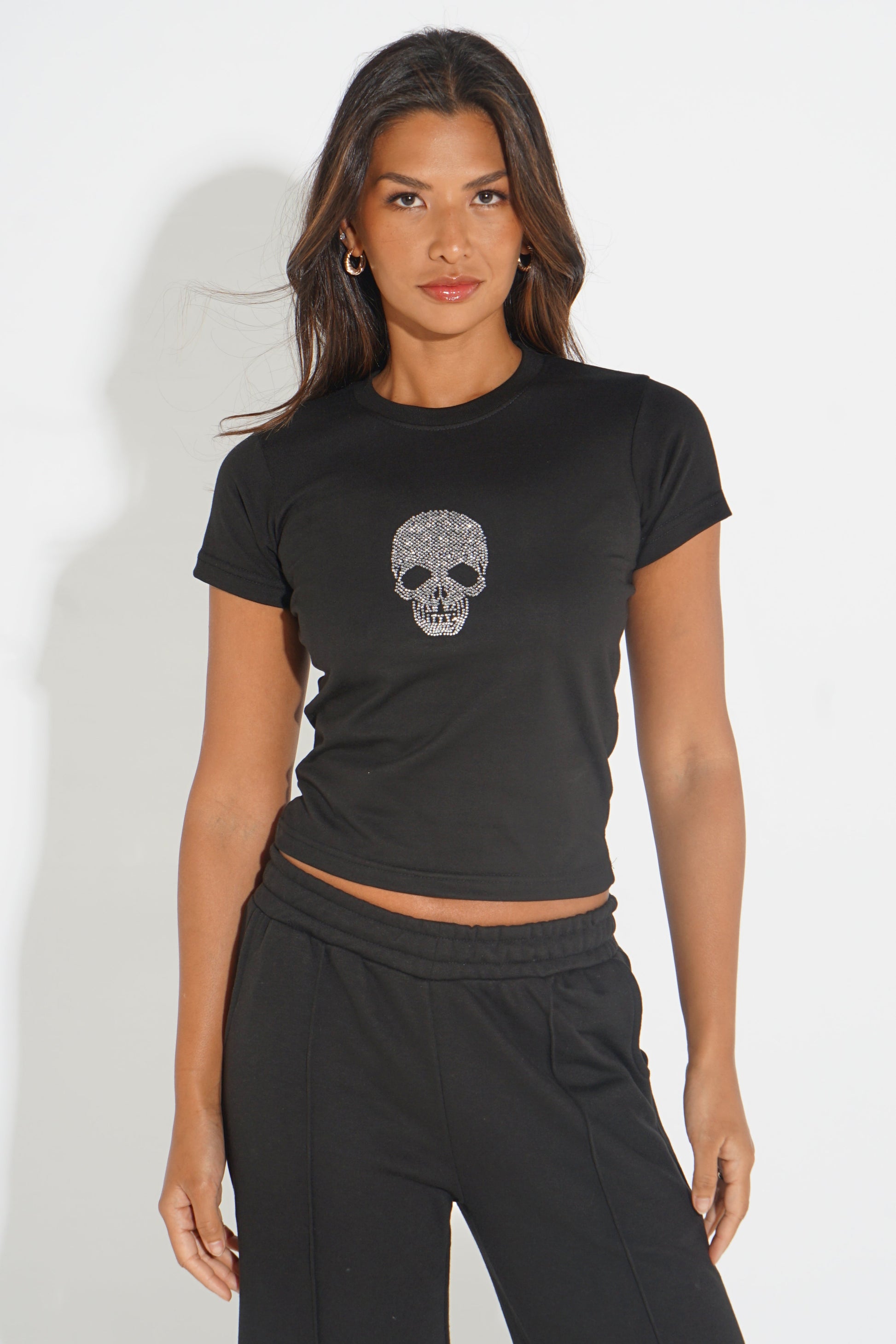 A woman modeling a black short-sleeve crop top with a silver glittery skull graphic on the chest, paired with black sweatpants, against a plain white studio background.