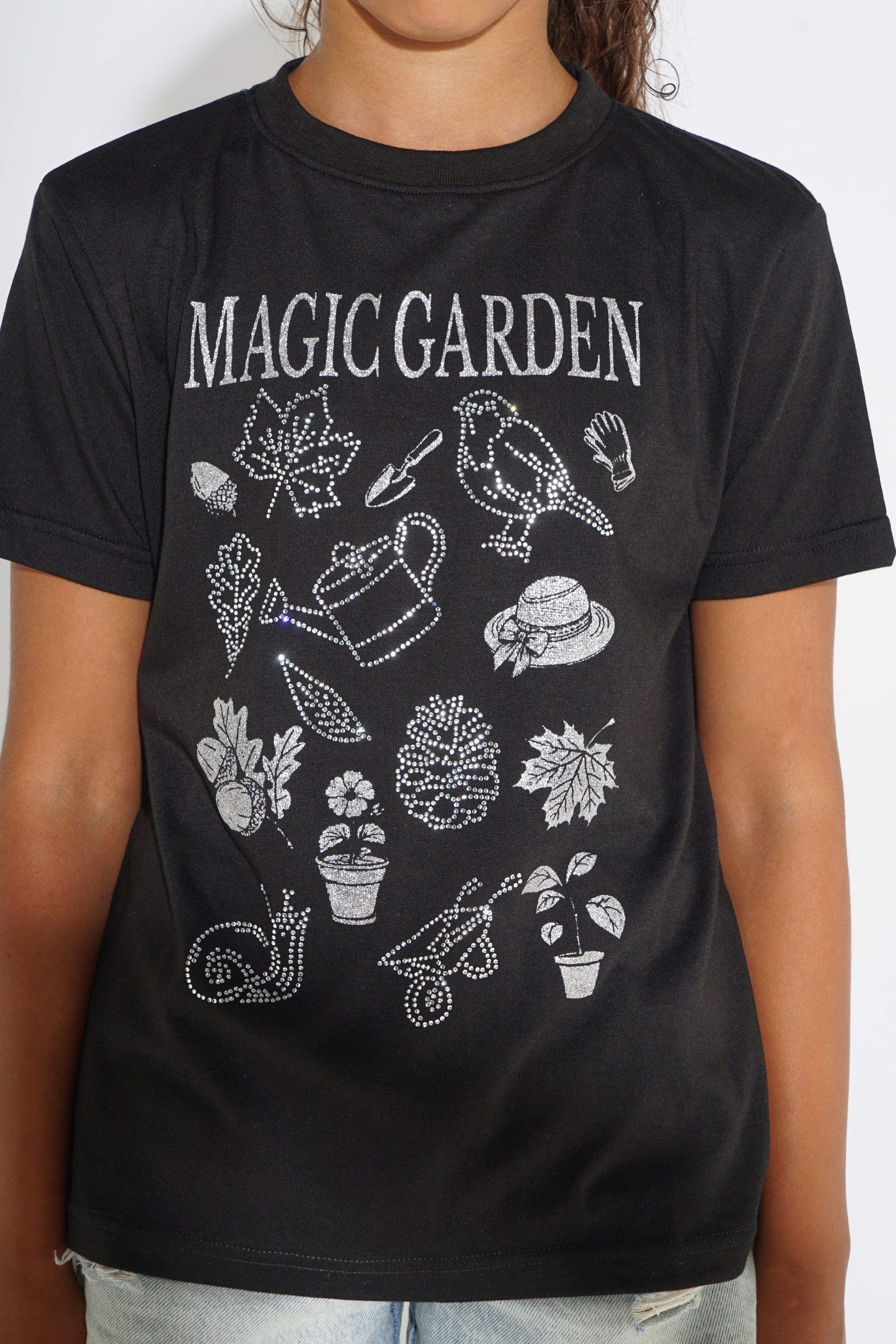 A black short-sleeve graphic t-shirt featuring the words 'MAGIC GARDEN' in white, with various white dotted illustrations of garden-themed items like a bird, watering can, plants, and leaves, worn by a model.