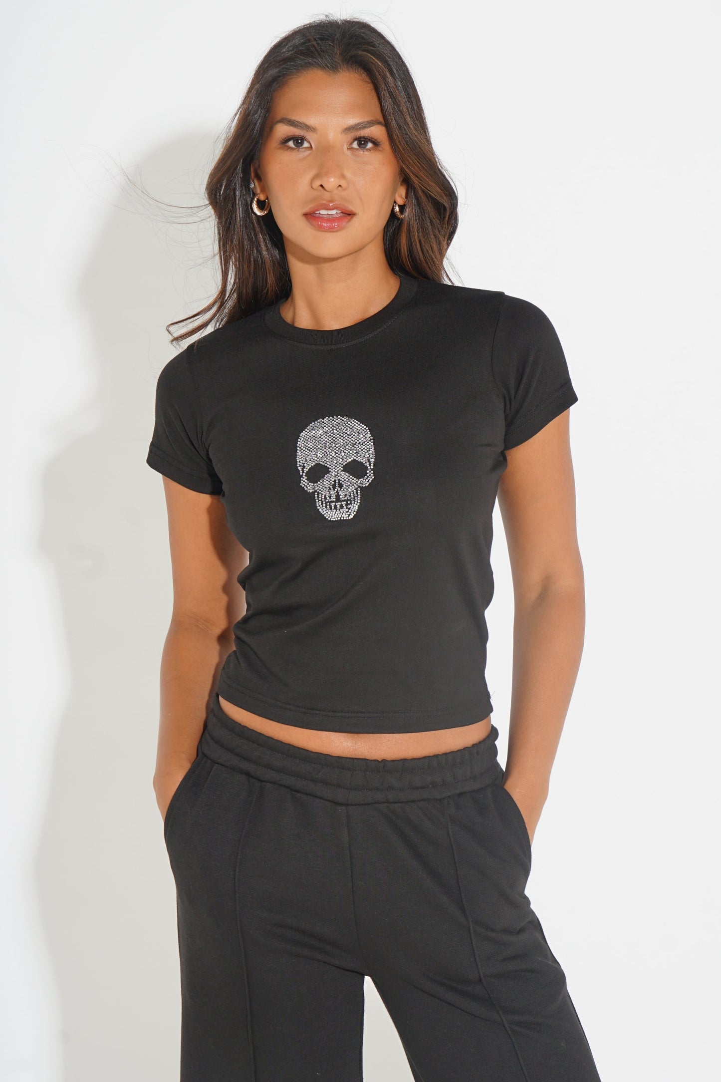 A woman modeling a black short-sleeve crop top with a gray skull graphic on the chest, paired with black sweatpants, against a plain white background.