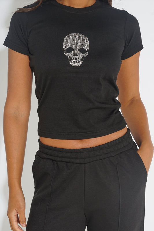 A woman wearing a black short-sleeve crop top with a silver skull graphic on the chest, paired with black sweatpants, shown against a plain light background.