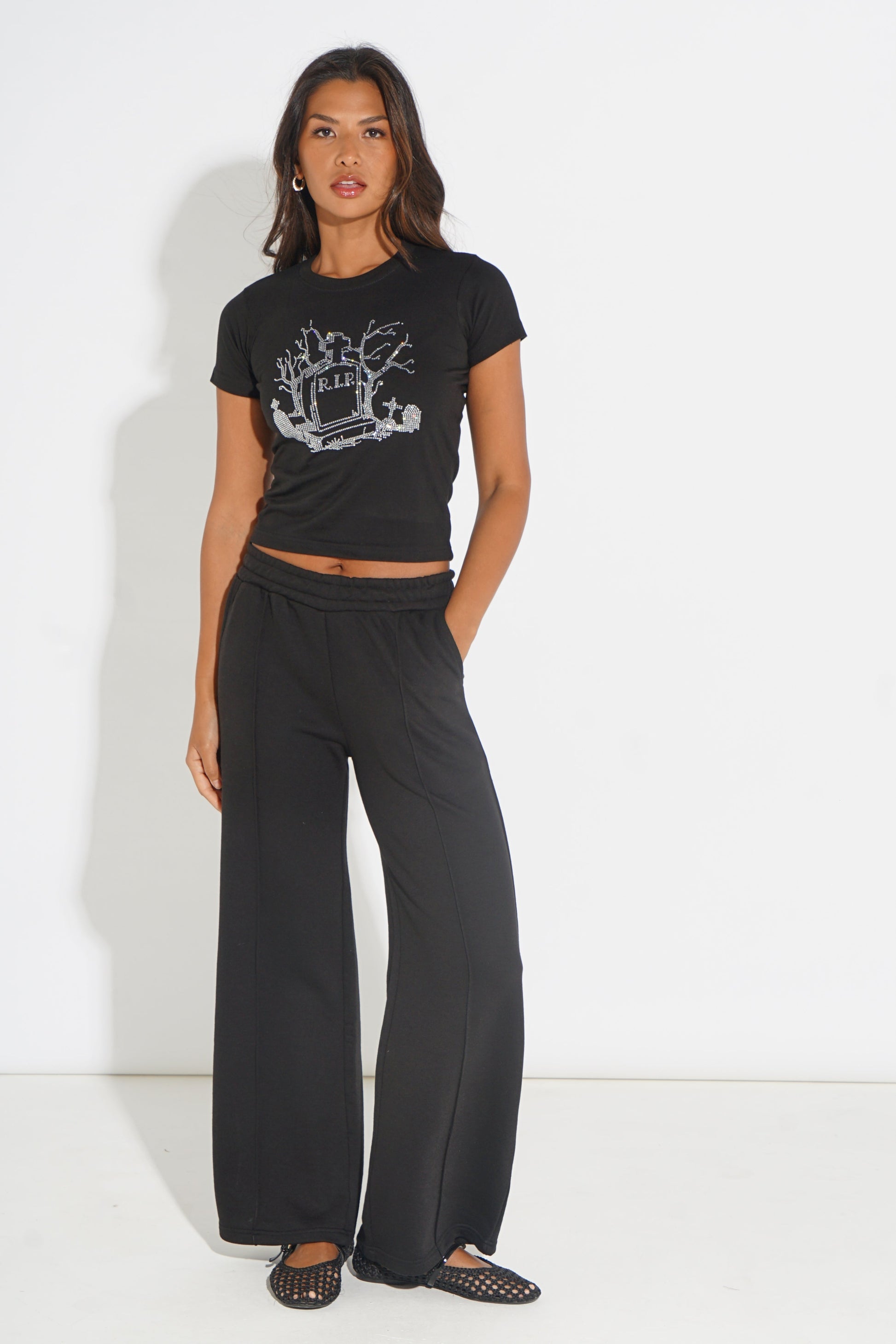 A woman modeling a black short-sleeve crop top with a white graphic print and black wide-leg pants, standing against a plain white studio background.