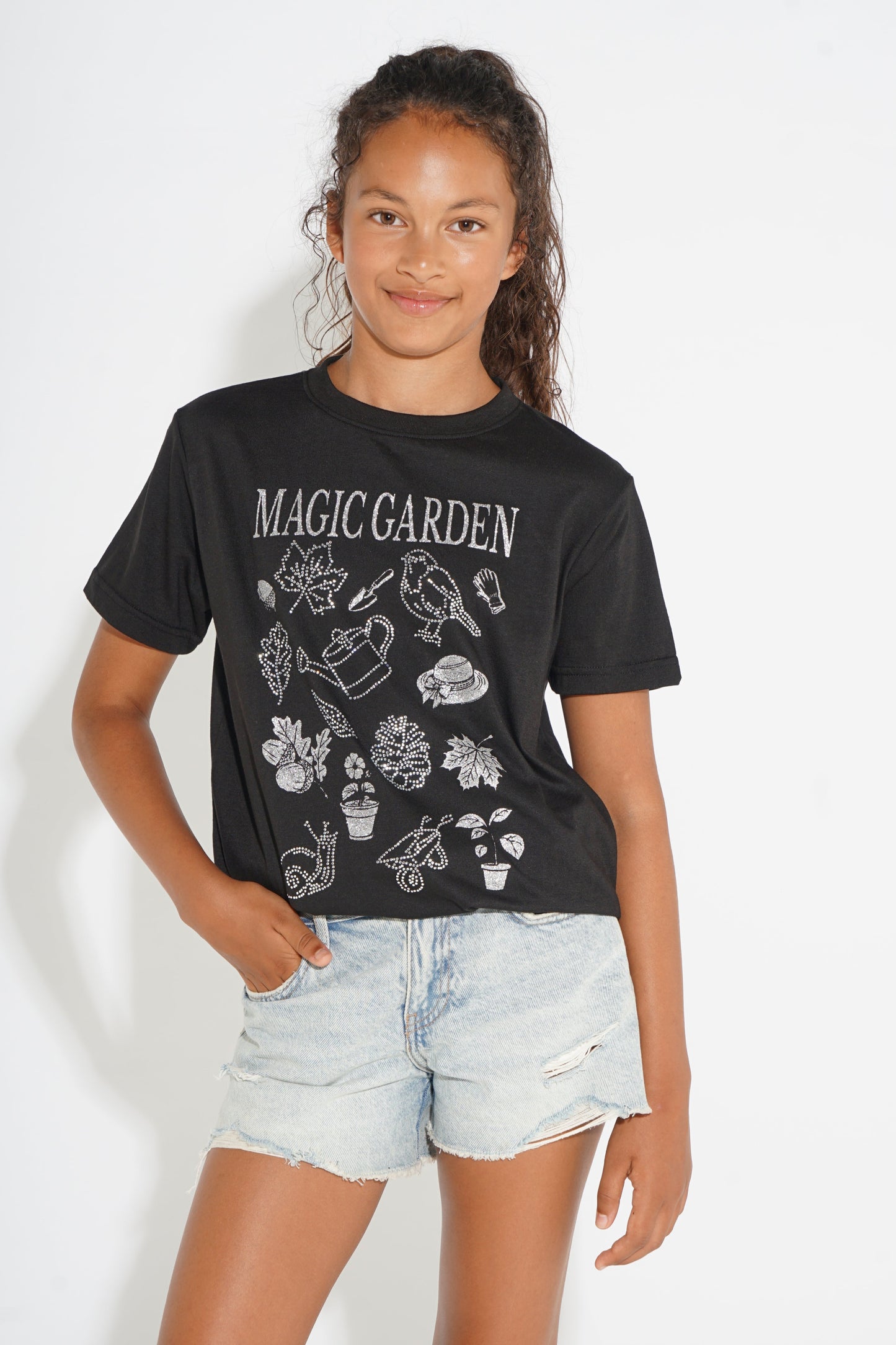 A young girl modeling a black short-sleeve graphic t-shirt with 'MAGIC GARDEN' and white gardening illustrations, paired with light-wash denim shorts against a white studio background.