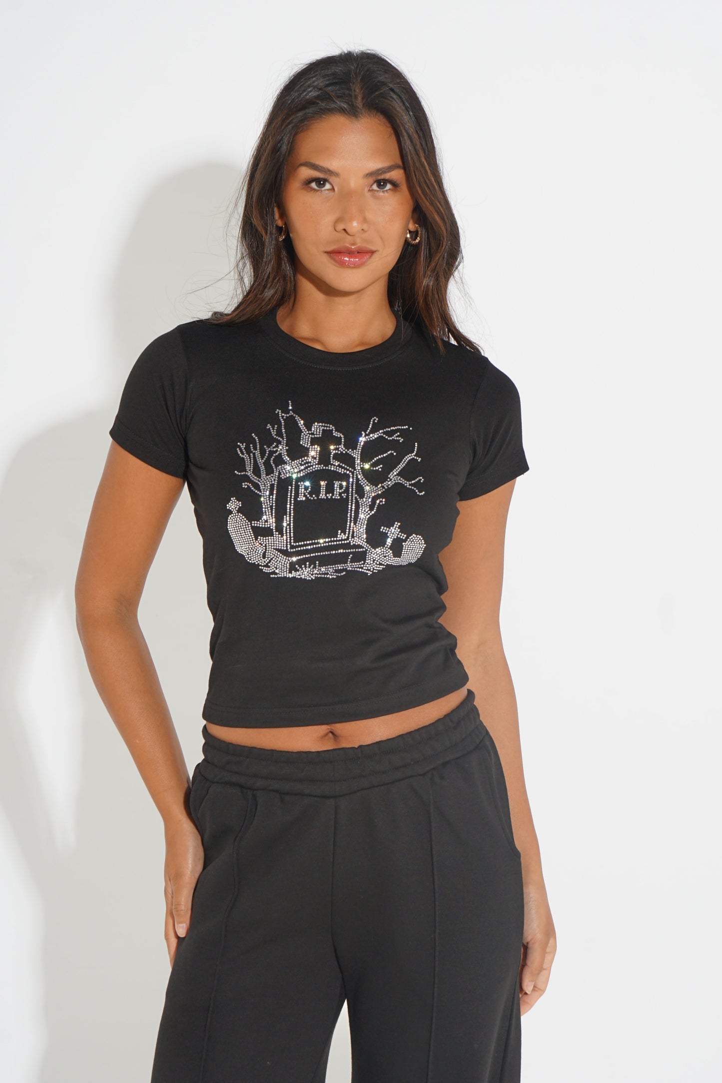A woman modeling a black short-sleeve crop top with a distressed white graphic on the chest, paired with black sweatpants, against a plain white studio background.