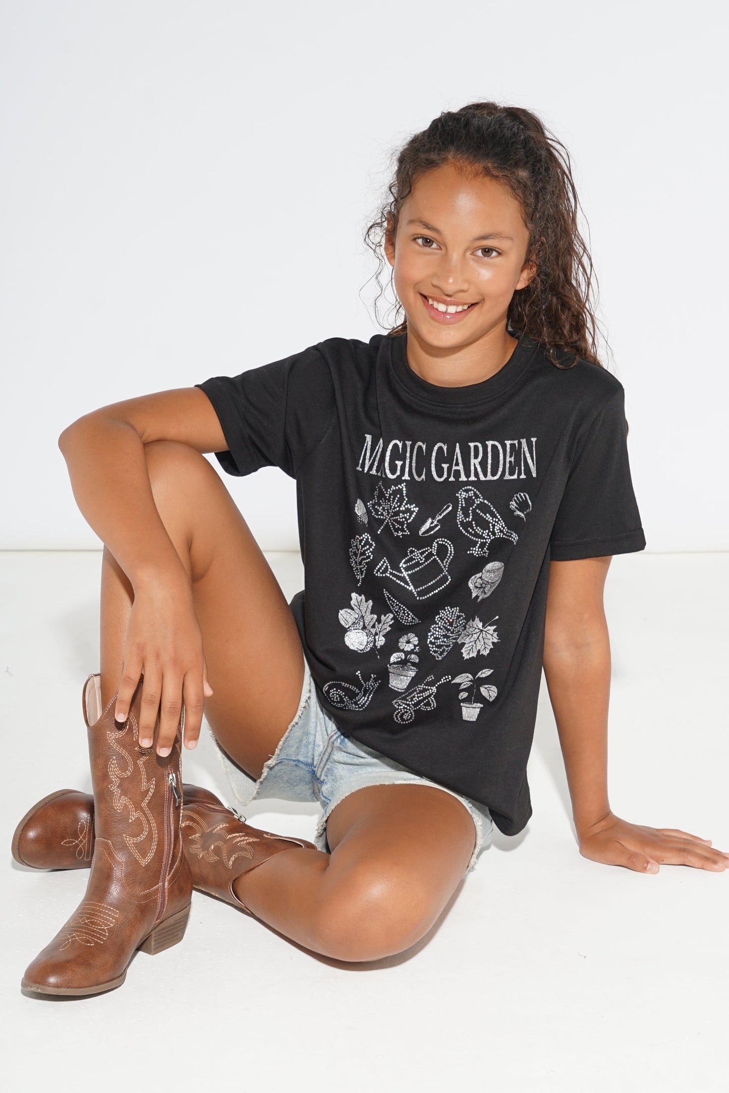 A young girl sitting on a white floor wearing a black graphic t-shirt with 'MAGIC GARDEN' printed in white, light-wash denim shorts, and brown cowboy boots.