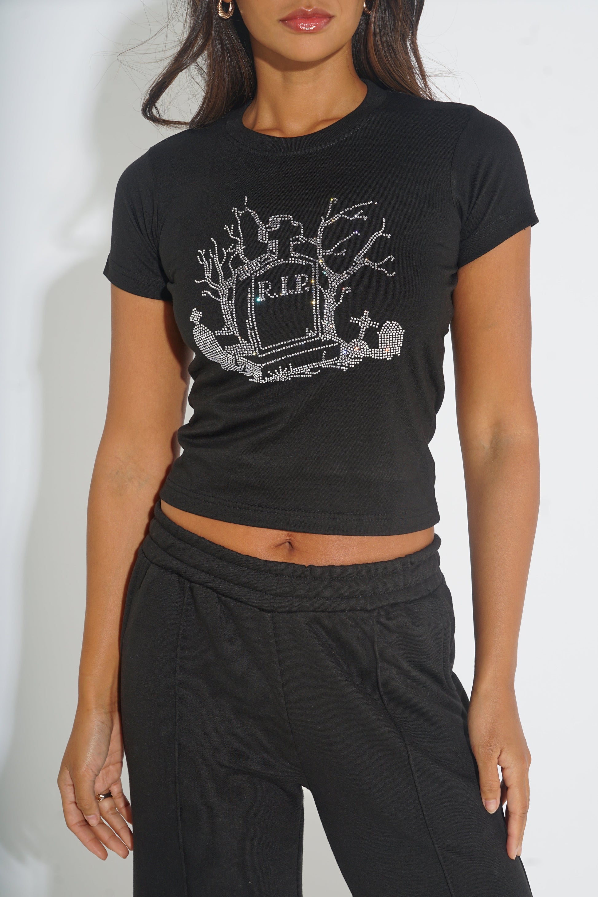 A woman wearing a black short-sleeve crop top with a silver glittery tombstone and tree graphic on the front, paired with black sweatpants, against a plain white background.